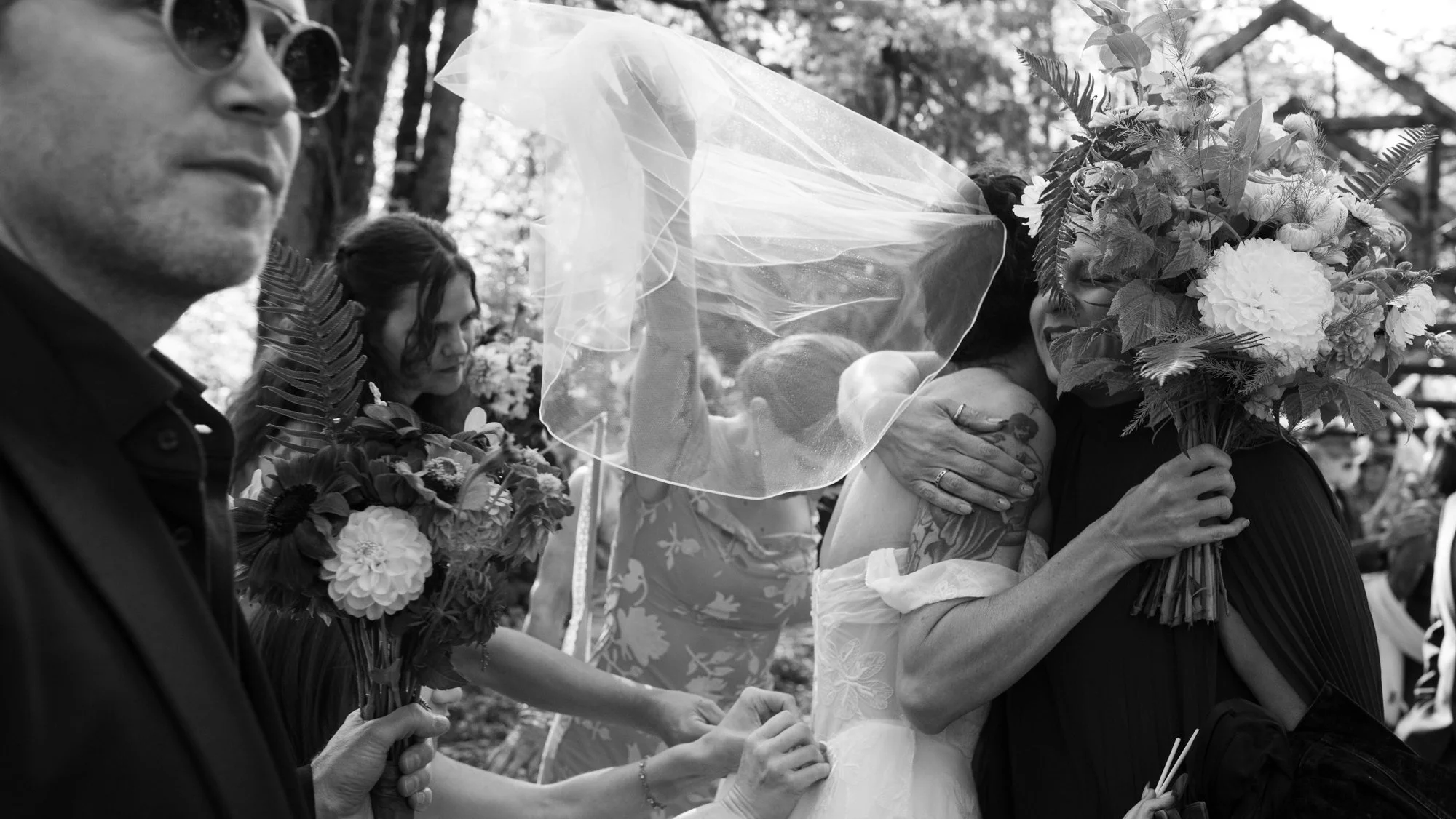 Black-and-white outdoor wedding scene with a veiled bride embracing a guest holding a large floral bouquet.