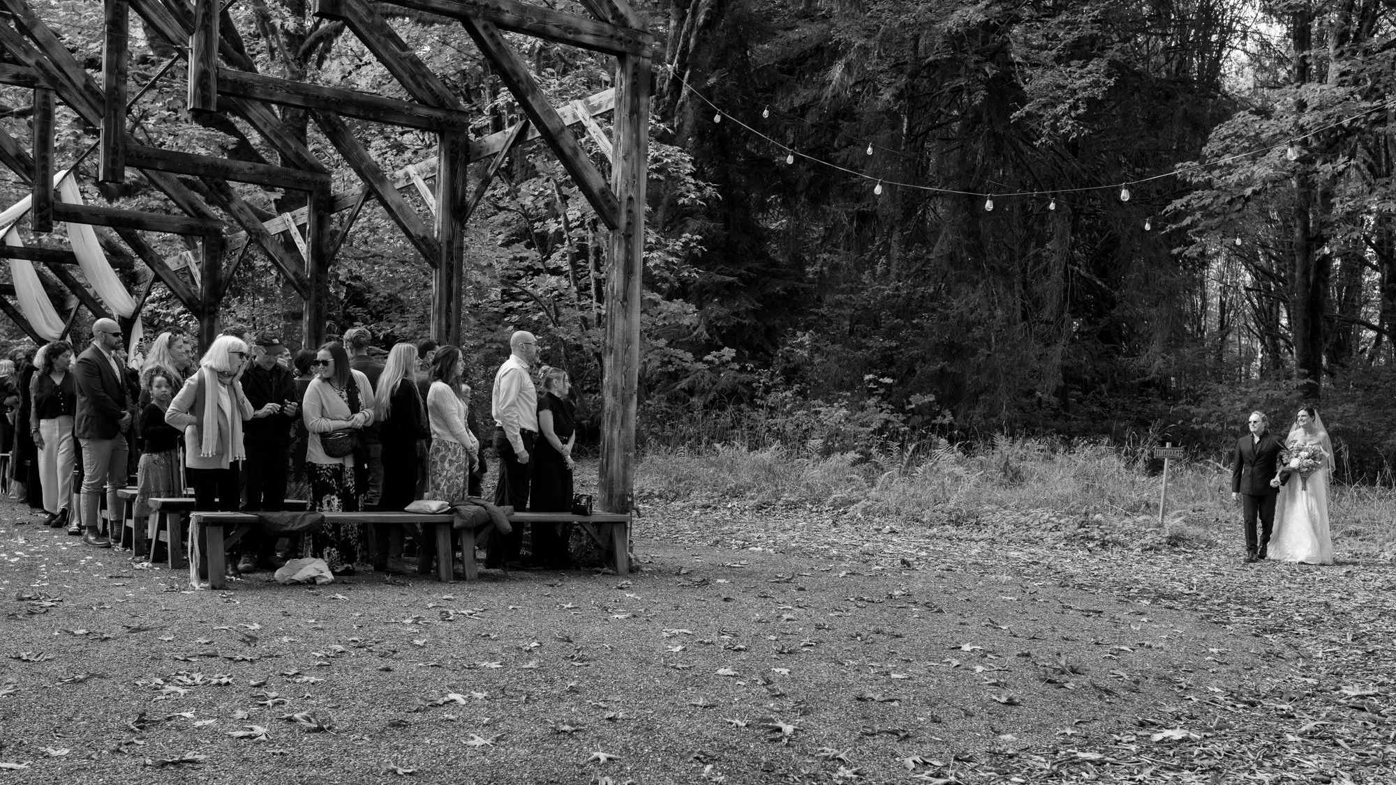 Bride and groom walk toward guests during a forest wedding ceremony beneath a rustic wooden canopy and string lights.