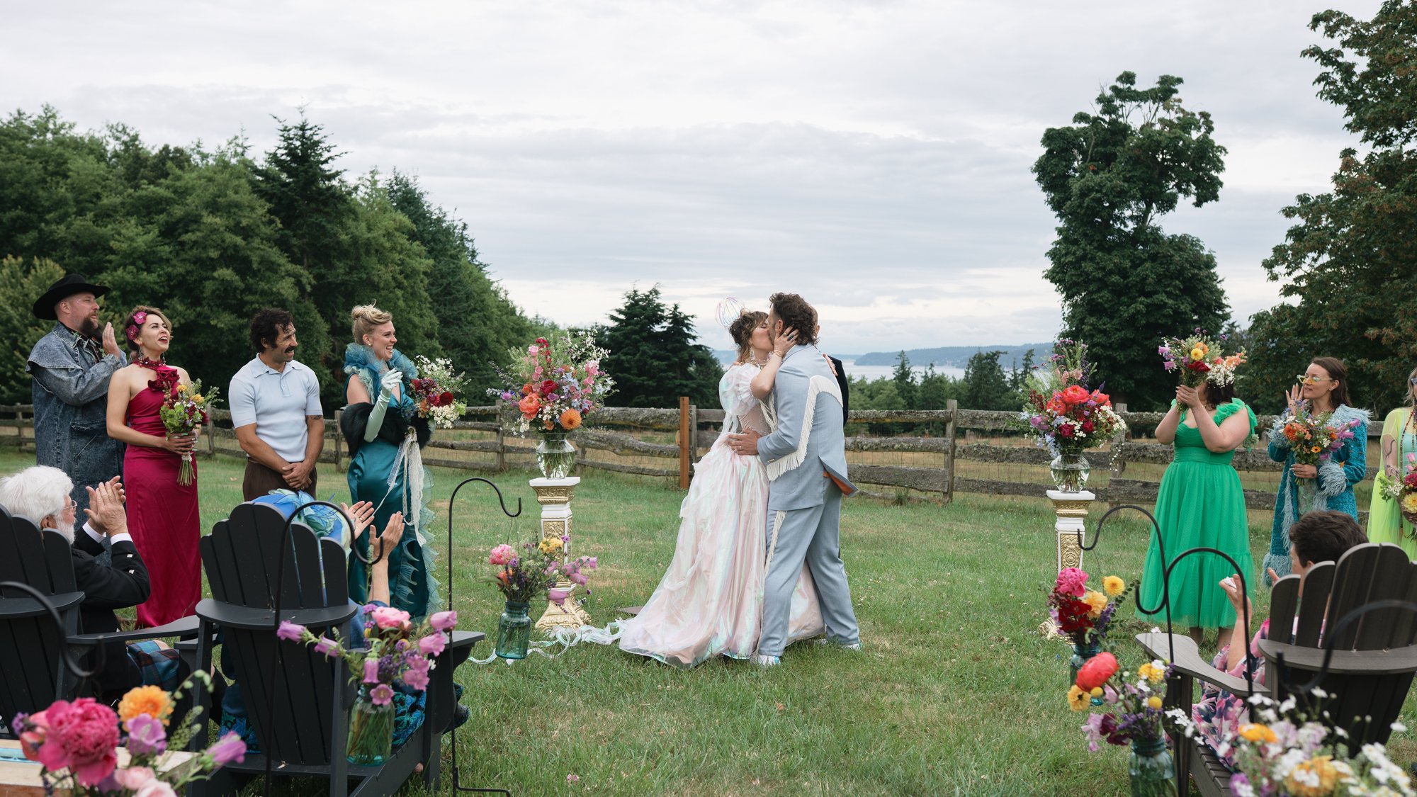 Newlywed couple kissing during an outdoor wedding ceremony surrounded by guests, floral arrangements, and wooded scenery.