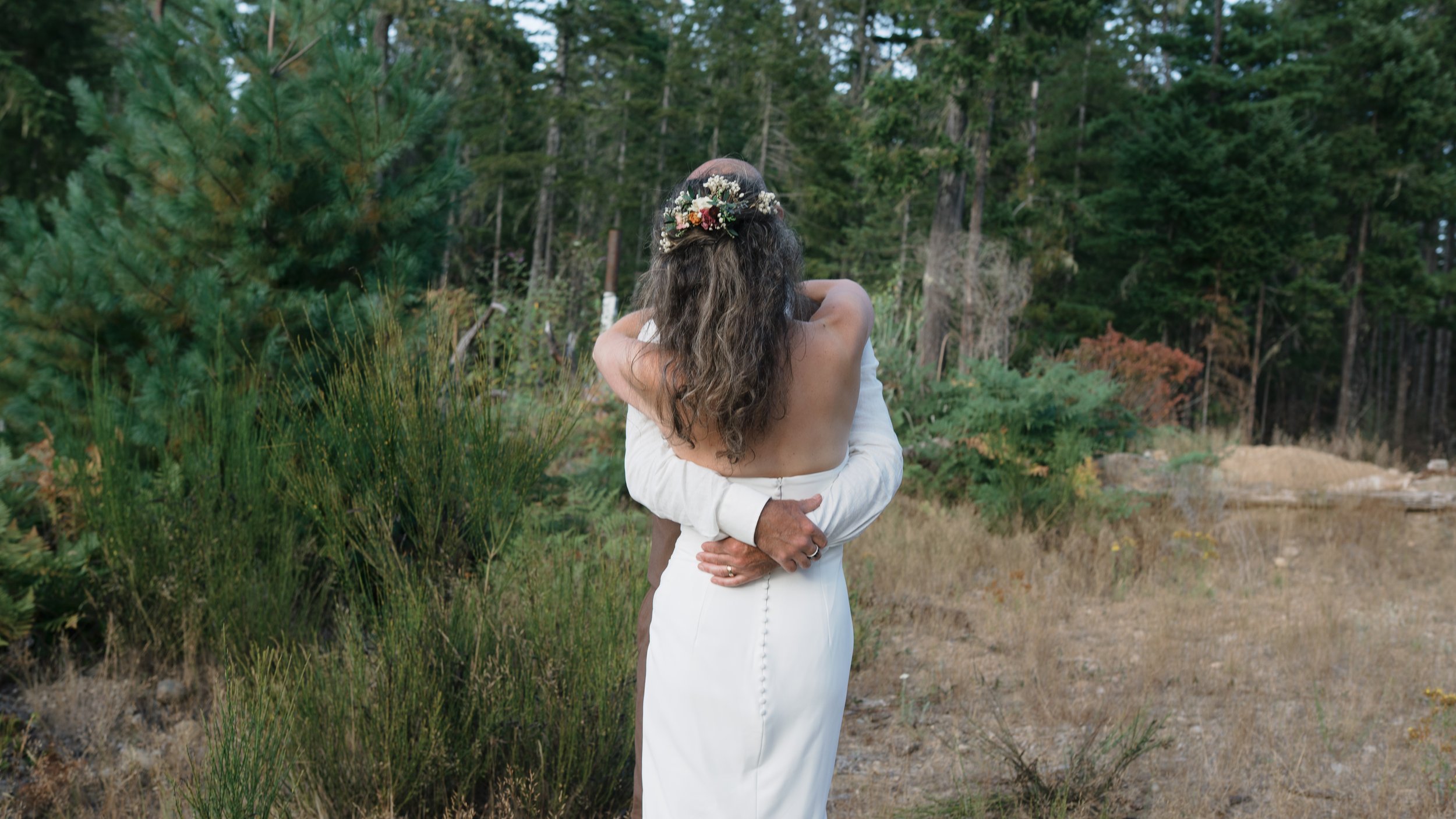 Bride in a white wedding dress and floral hairpiece embraces a groom in a forest clearing.