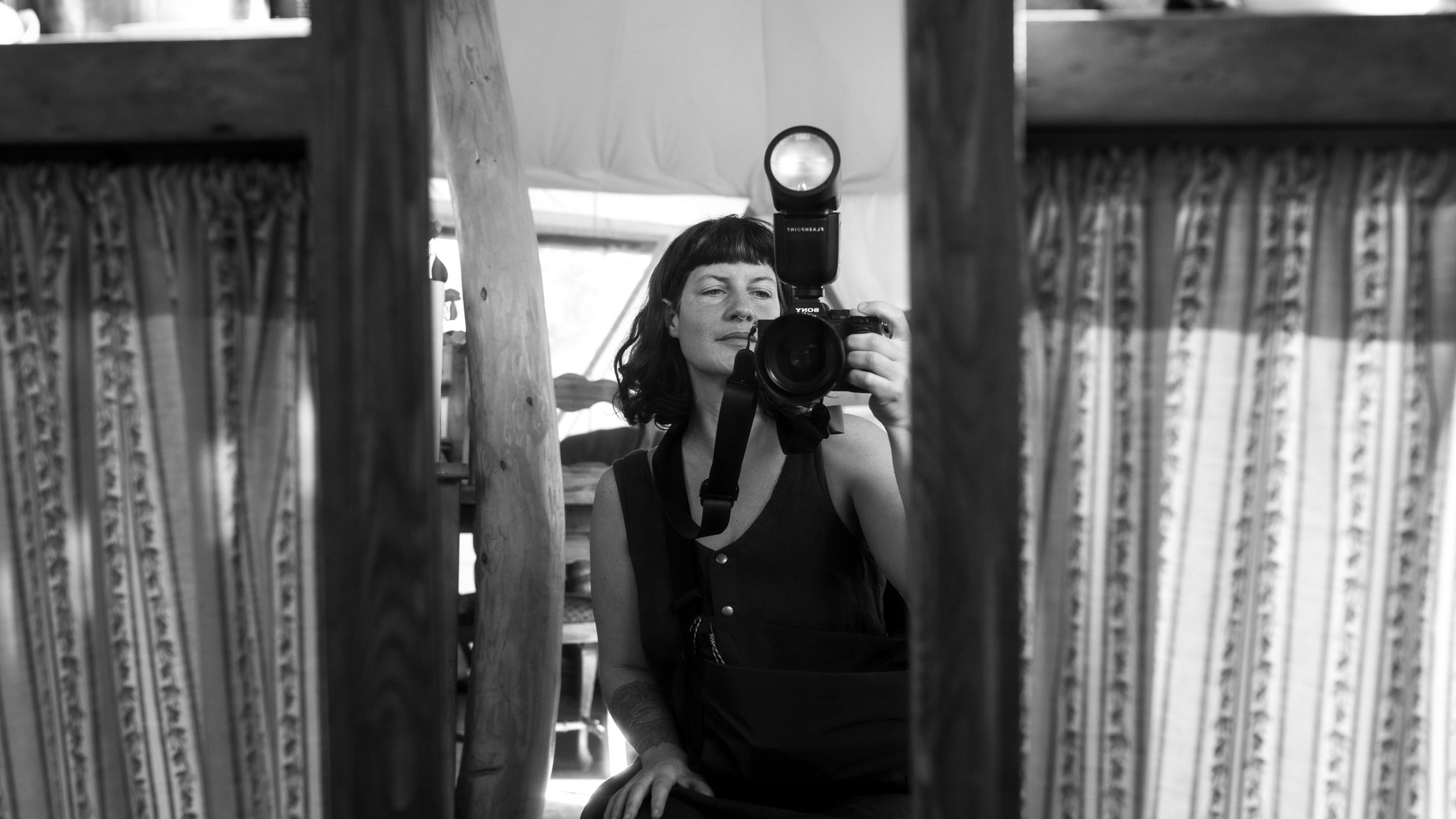 Black-and-white mirror selfie of a woman holding a Sony camera with an external flash in a rustic room