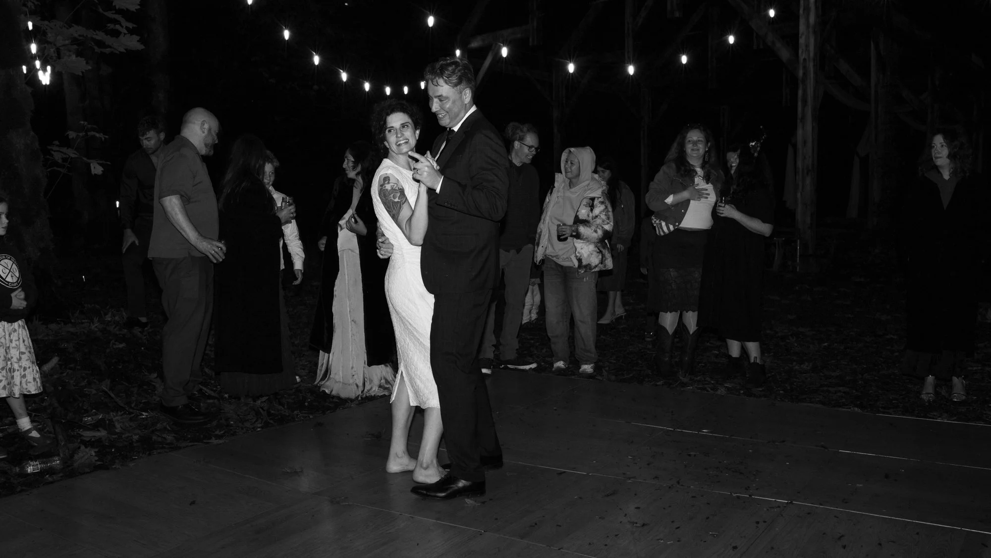 Black-and-white photo of a couple dancing on a wooden floor at an outdoor nighttime wedding reception, surrounded by guests and string lights.