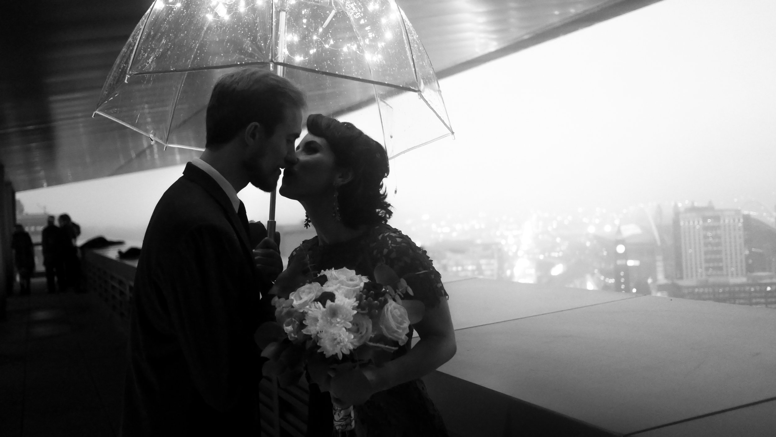 Black-and-white photo of a couple kissing under a clear umbrella while the woman holds a bouquet of flowers.