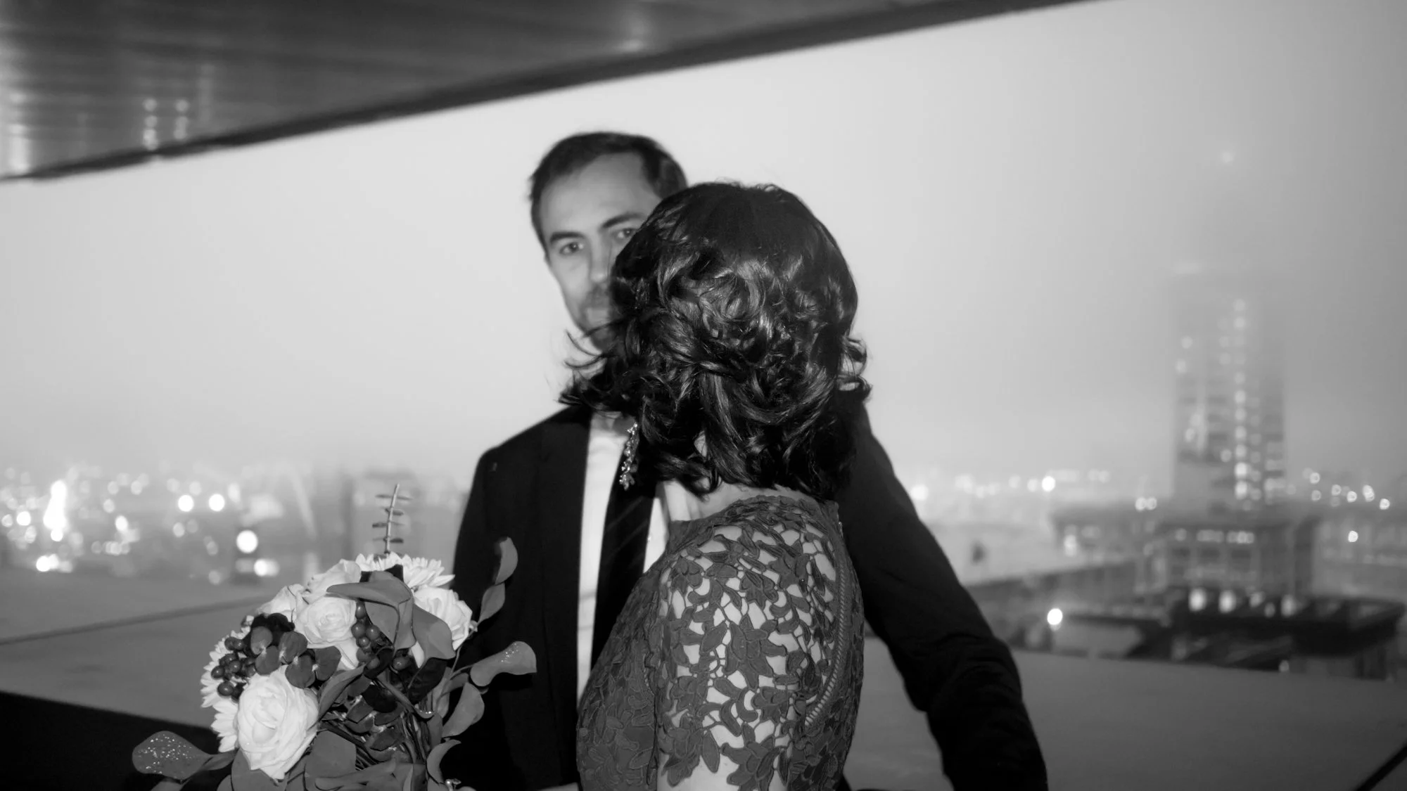 Black-and-white photo of a formally dressed couple embracing on a rooftop at night, with a bouquet and city skyline in the background.