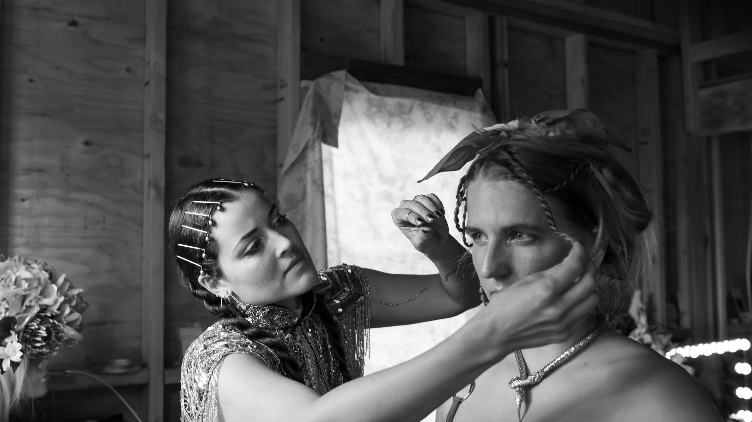 Black-and-white photo of a stylist applying theatrical makeup to a costumed performer in a rustic wooden dressing room