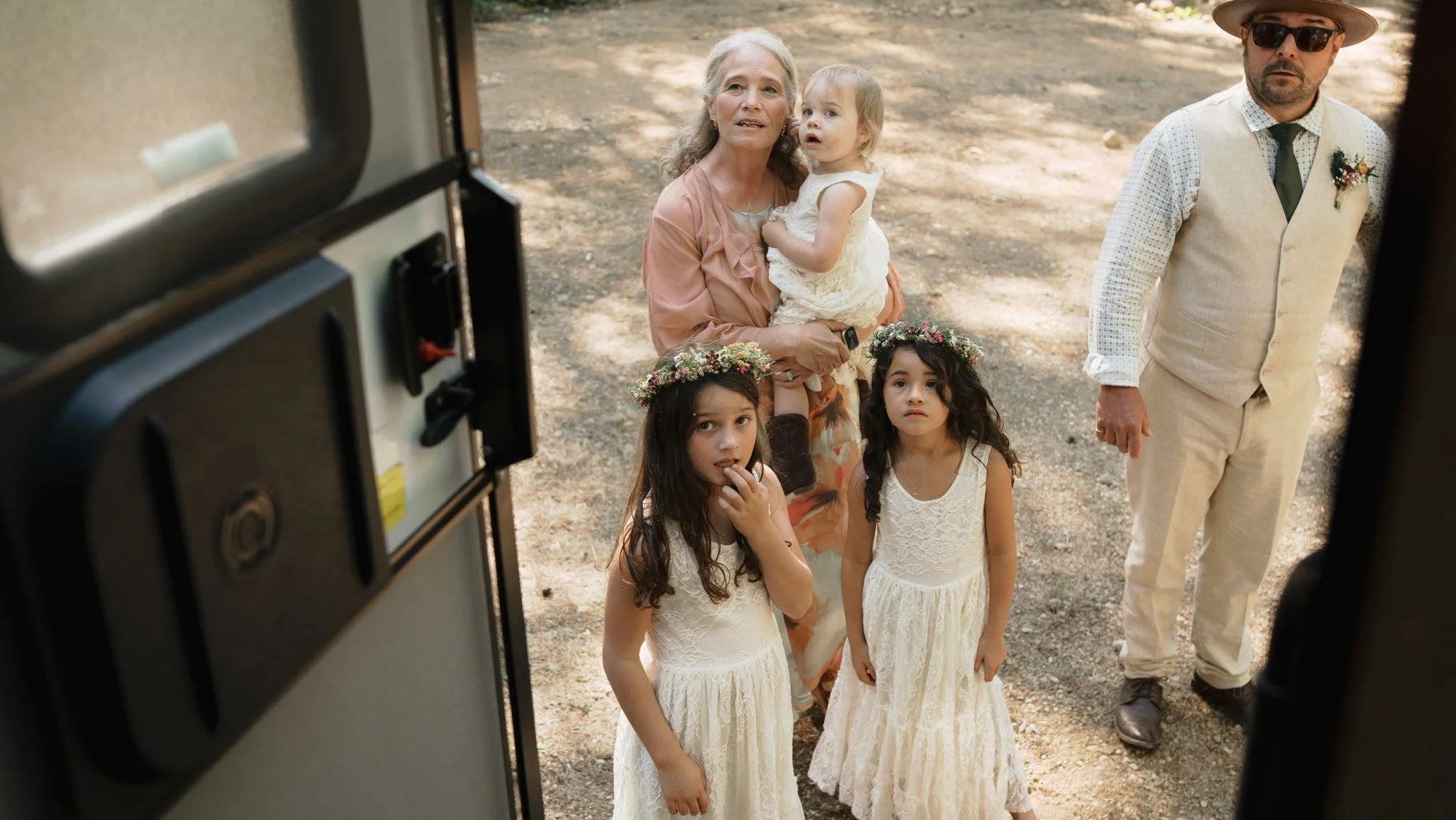 Family in wedding attire gathered outside a vehicle, with two flower-crowned flower girls and a toddler in a white dress