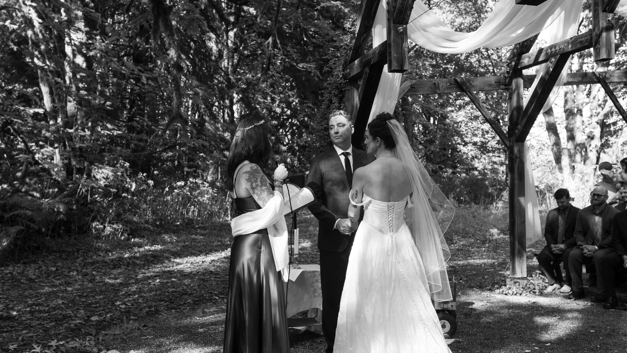 Black-and-white outdoor wedding ceremony in a forest, with a bride and groom under a draped wooden arbor as an officiant speaks.