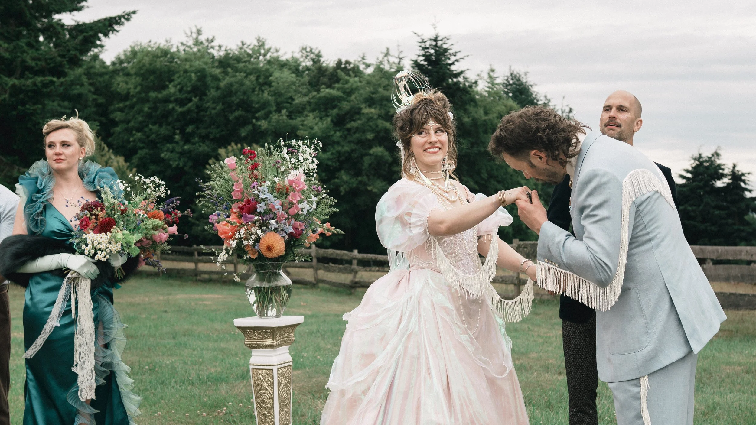 Wedding party in colorful formal attire holding flower bouquets outdoors as a bride dances with two men in a garden.