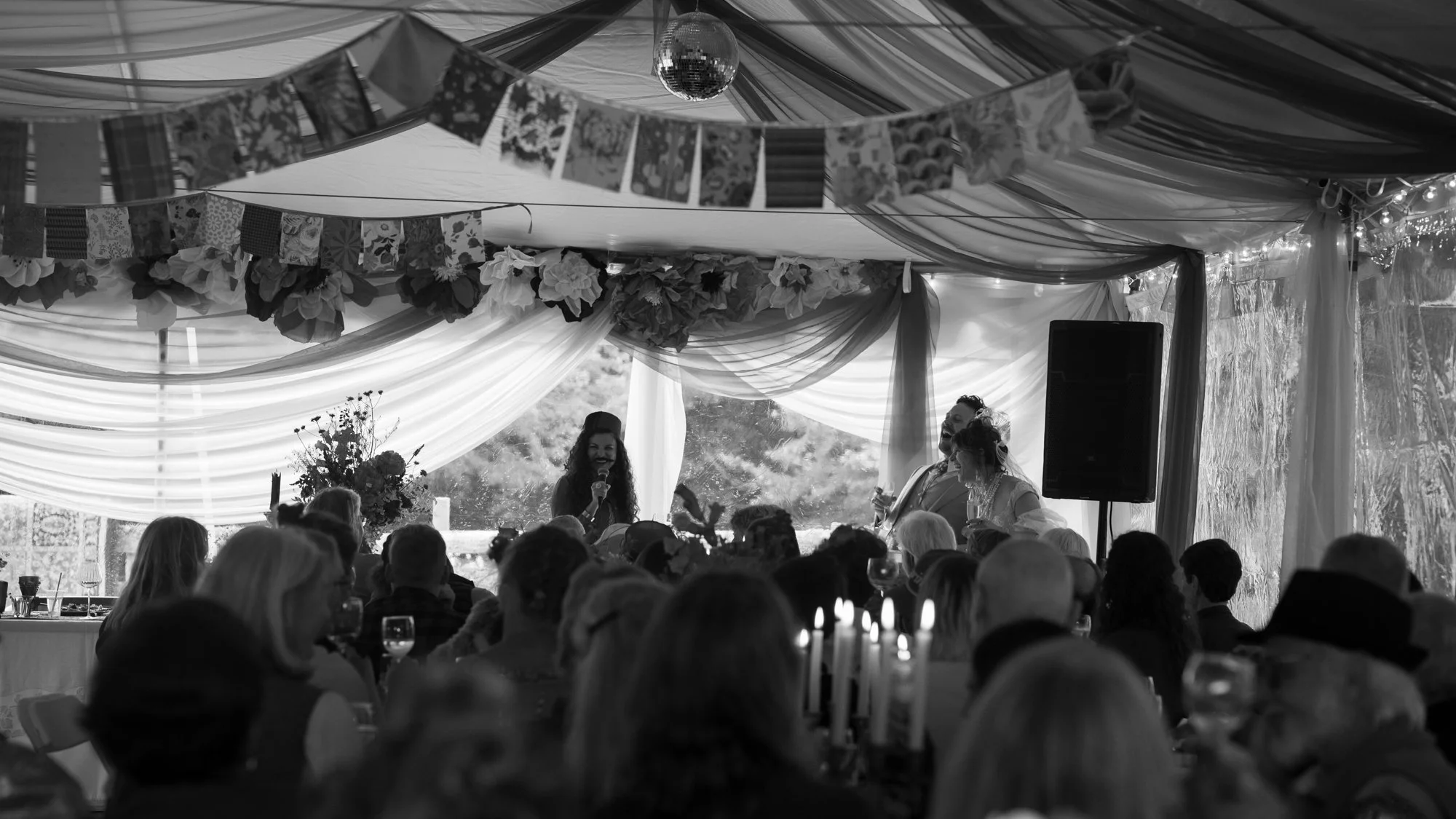 Guests attend a wedding reception under a decorated tent as a woman gives a speech beside a bride and groom.