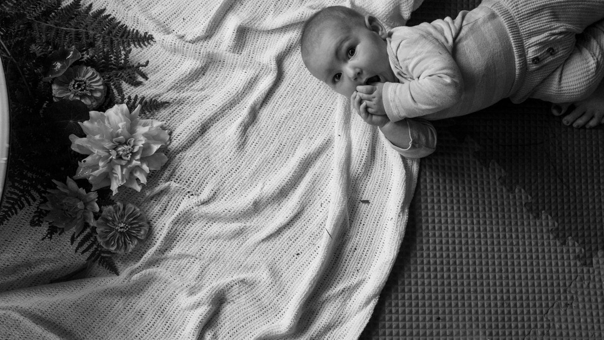 Black-and-white photo of a baby lying on a knitted blanket beside flowers.