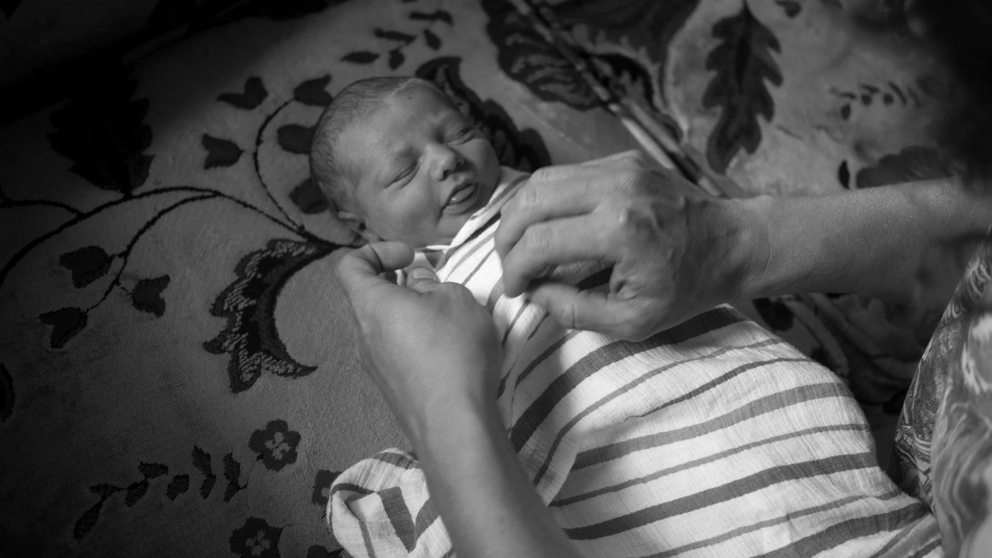 Black-and-white photo of an adult gently swaddling a sleeping newborn baby in a striped blanket.