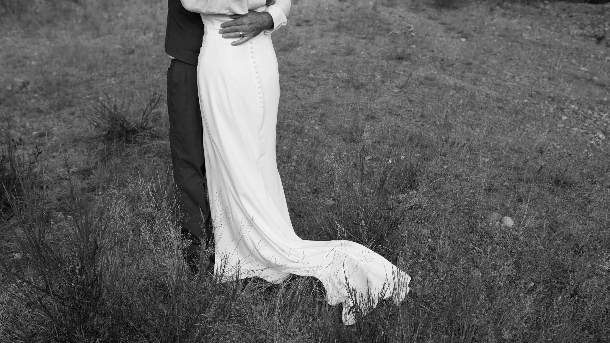Black-and-white wedding photograph showing a bride in a long button-back gown embracing the groom in a grassy field