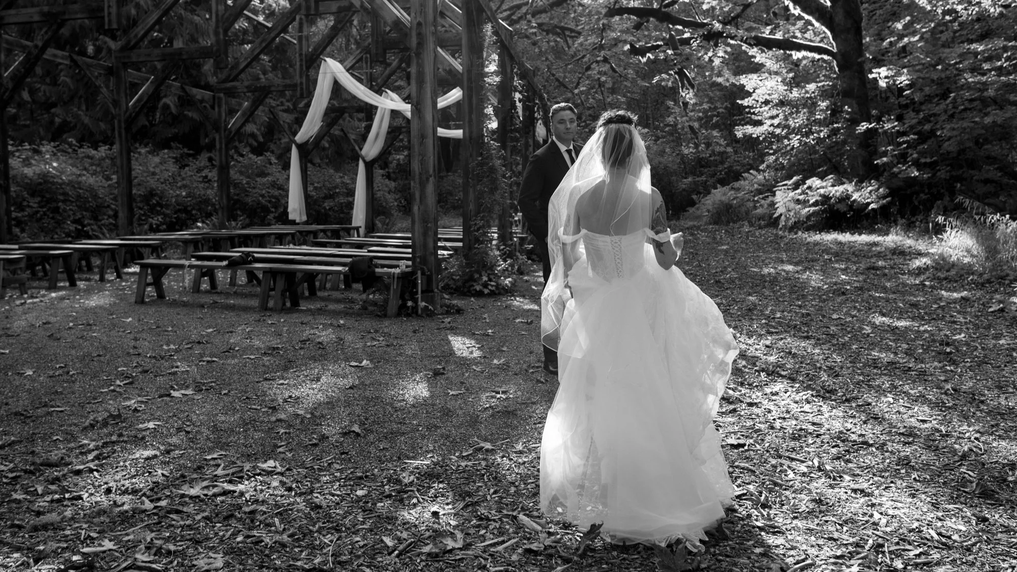 Black-and-white photo of a bride and groom at a woodland wedding venue with draped ceremony beams and wooden picnic benches.