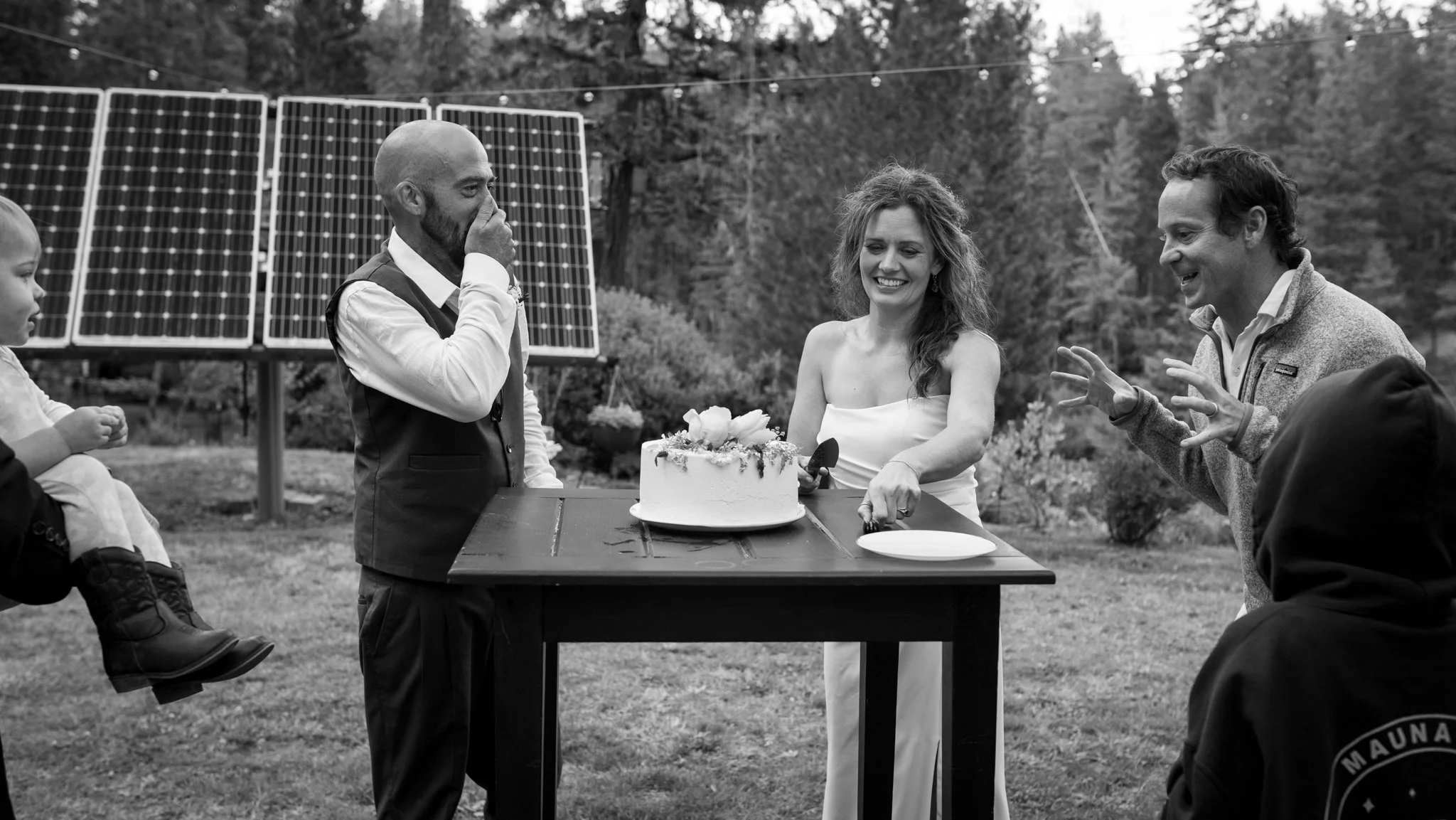 Wedding guests gather around a cake outdoors beside solar panels.