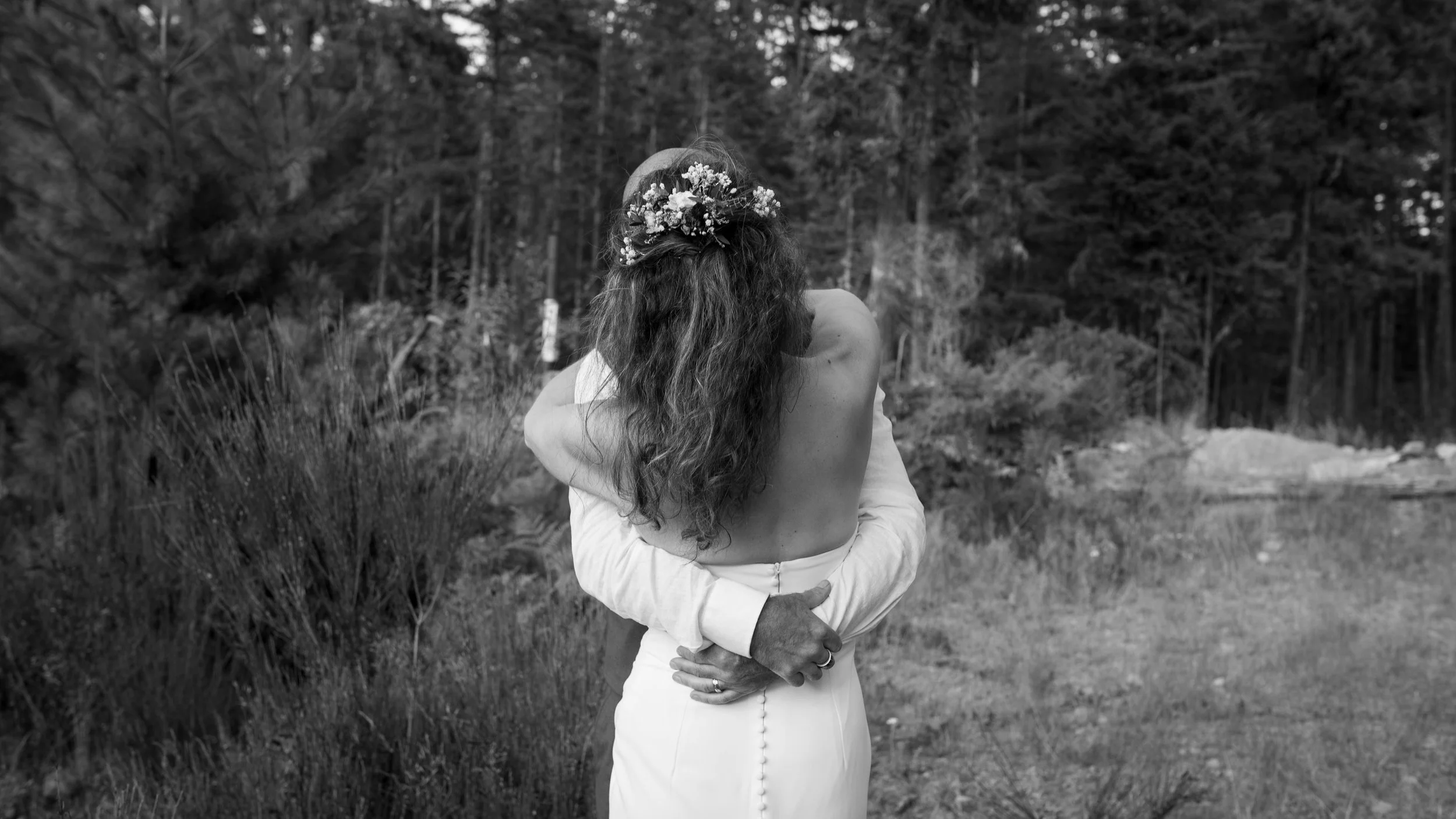 Black-and-white photo of a bride with long hair and a floral hairpiece embracing her partner in a forest.