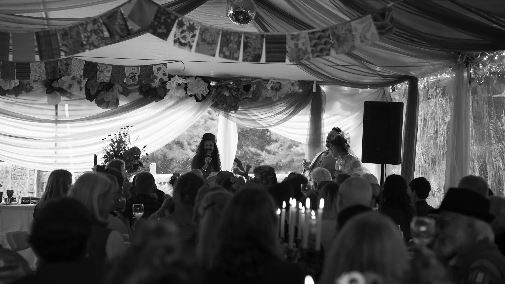 Black-and-white wedding reception with a guest giving a speech beneath floral decorations and draped fabric, surrounded by seated guests.
