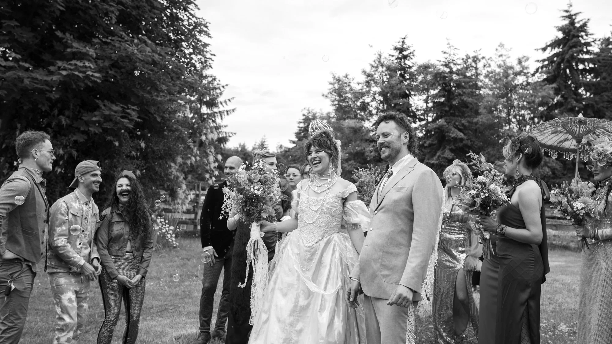 Bride and groom laughing with wedding guests outdoors as bubbles float around them