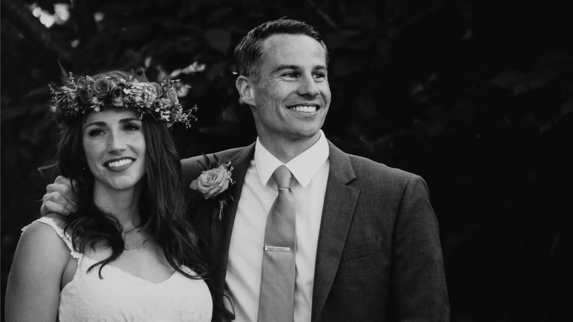 Black-and-white wedding portrait of a smiling bride wearing a floral crown and groom in a suit with a boutonniere