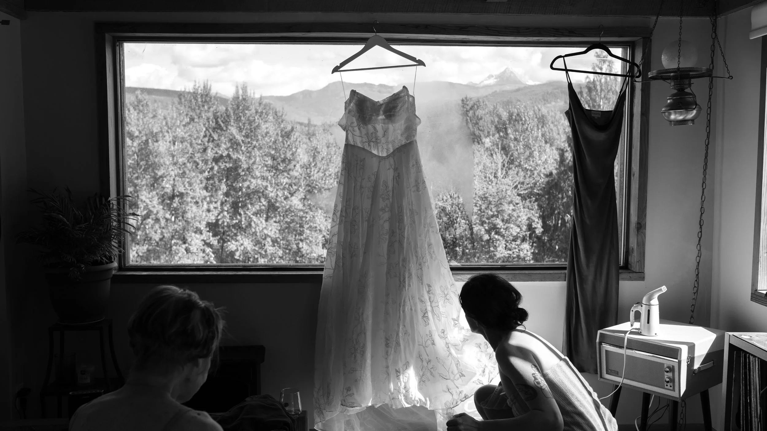 Black-and-white photo of a floral dress hanging in front of a mountain view, with two people seated nearby.