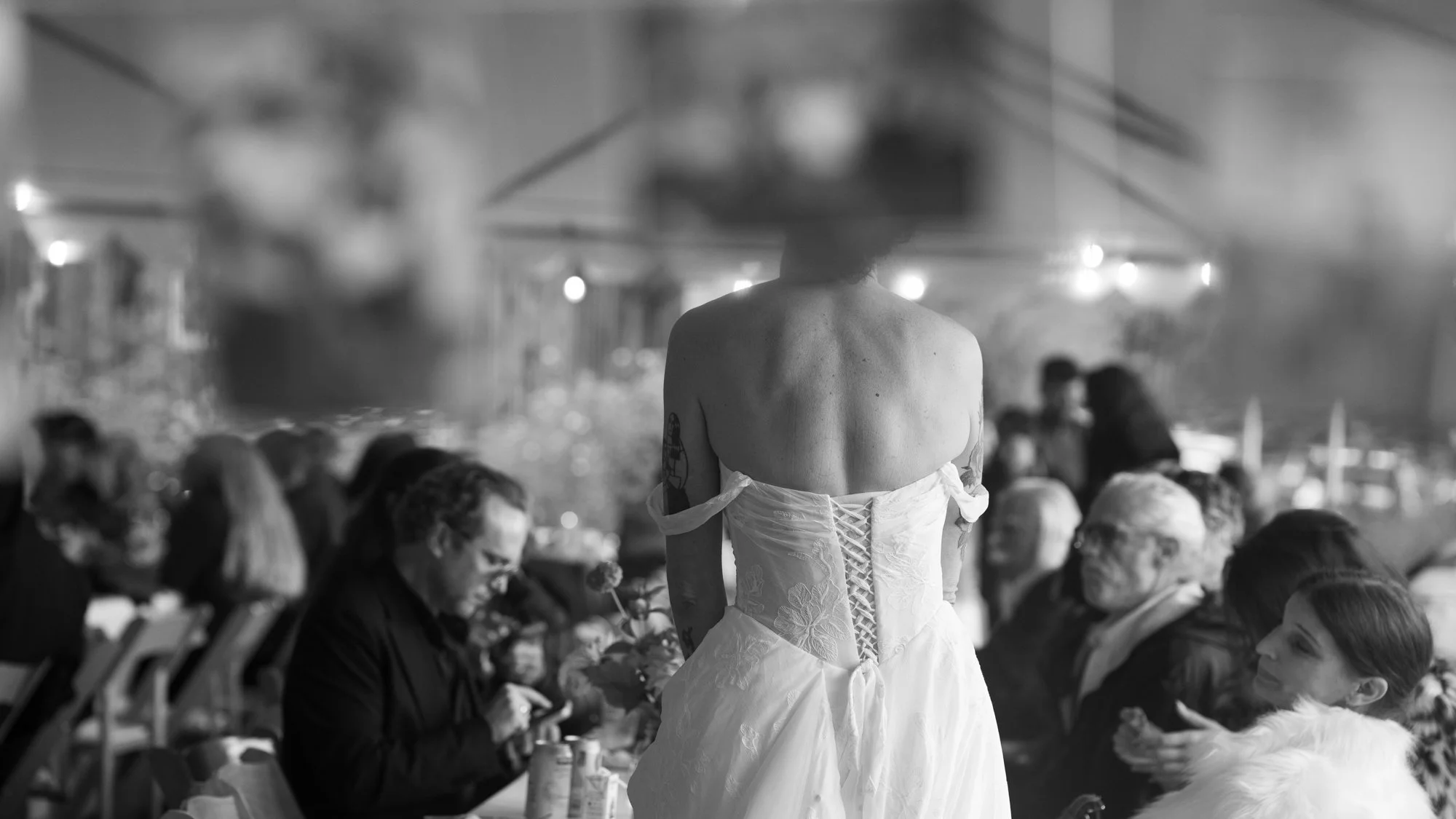 Tattooed woman wearing a strapless white corset-style wedding dress at a crowded reception