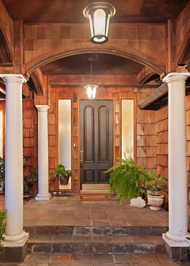 Welcoming wood-shingled porch with white columns, black front door, lantern lights, and potted ferns