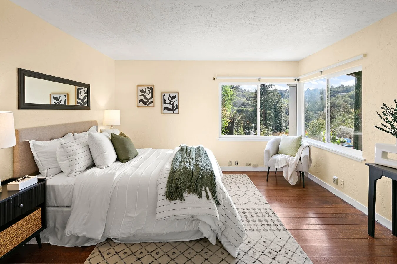 Bright neutral-toned bedroom with a neatly made bed, green throw blanket, armchair, large windows, and hardwood floors overlooking trees.