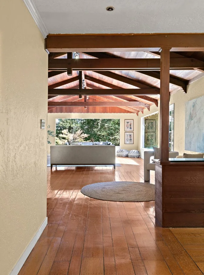 Sunlit open-plan living room with exposed wood beams, hardwood floors, gray sofa, round rug, and large windows overlooking trees.
