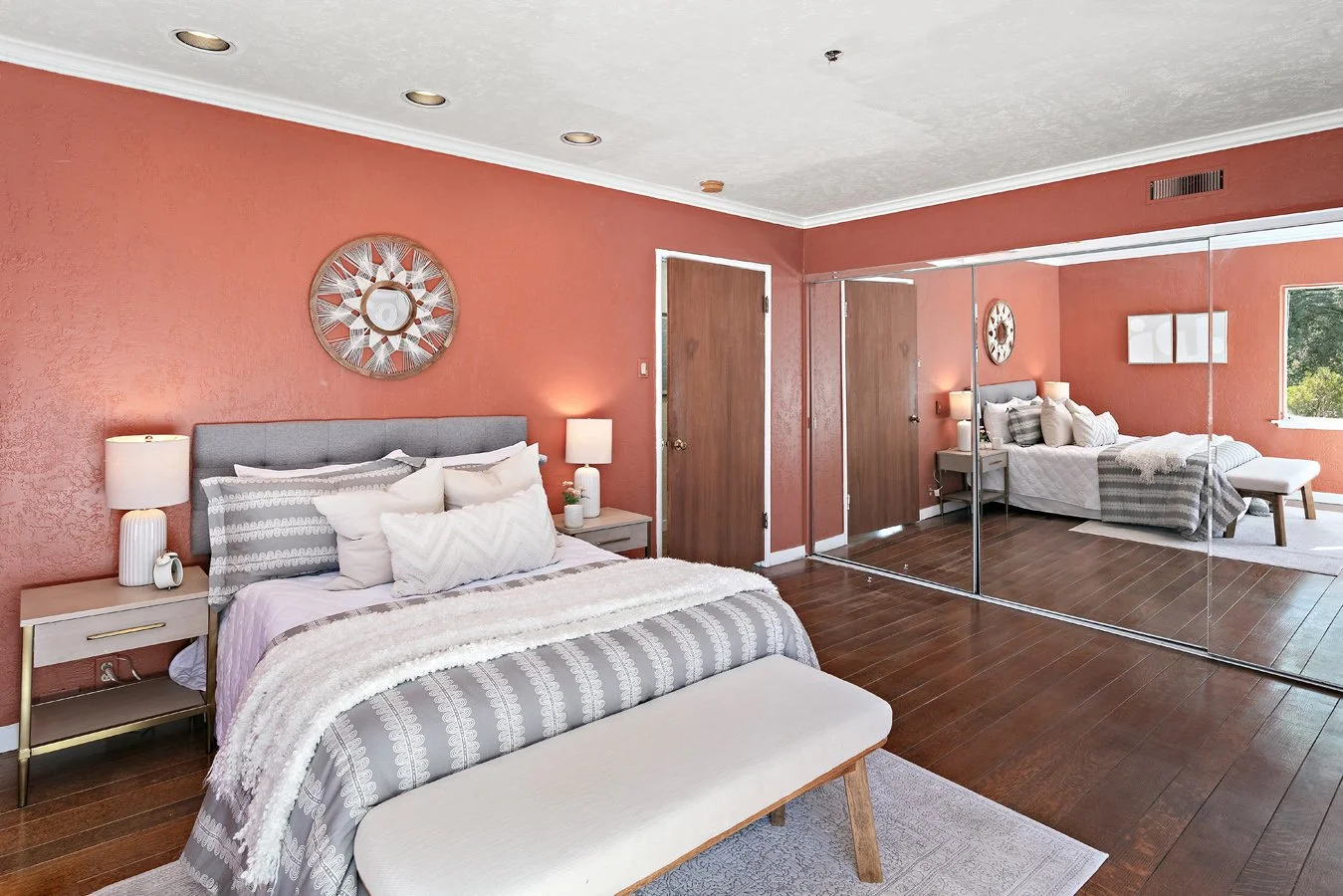 Stylish coral bedroom with a gray upholstered bed, patterned bedding, large mirrored closet doors, and a cushioned bench.