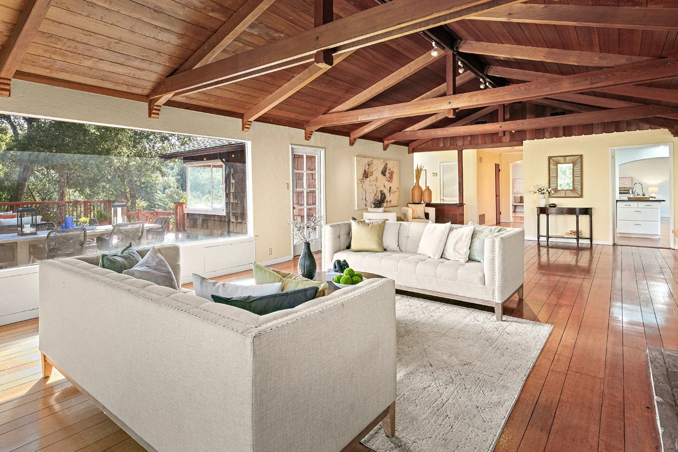 Bright open-concept living room with white sofas, vaulted exposed wood-beam ceiling, hardwood floors, and large windows overlooking a wooded deck.