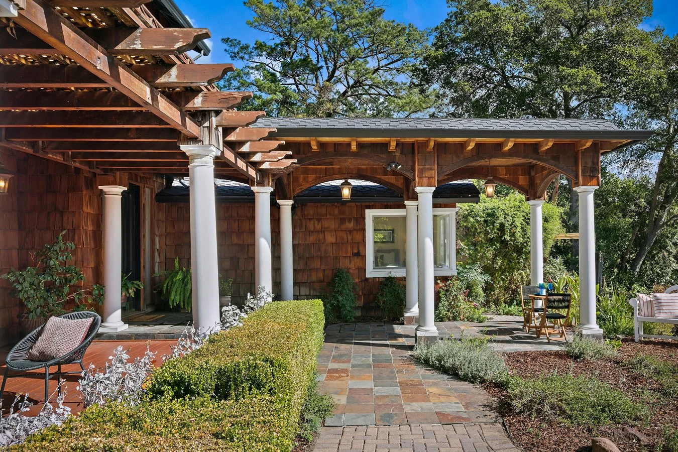 Wood-shingled home with a covered patio, white columns, landscaped courtyard, and outdoor seating.