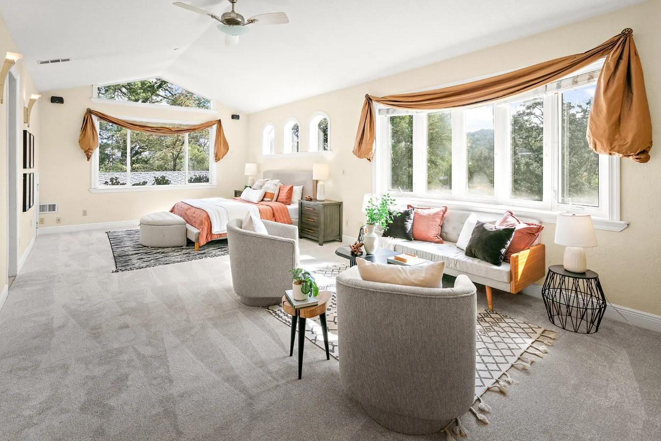 Bright bedroom sitting area with a bed, gray armchairs, sofa, large windows, and gold drapery