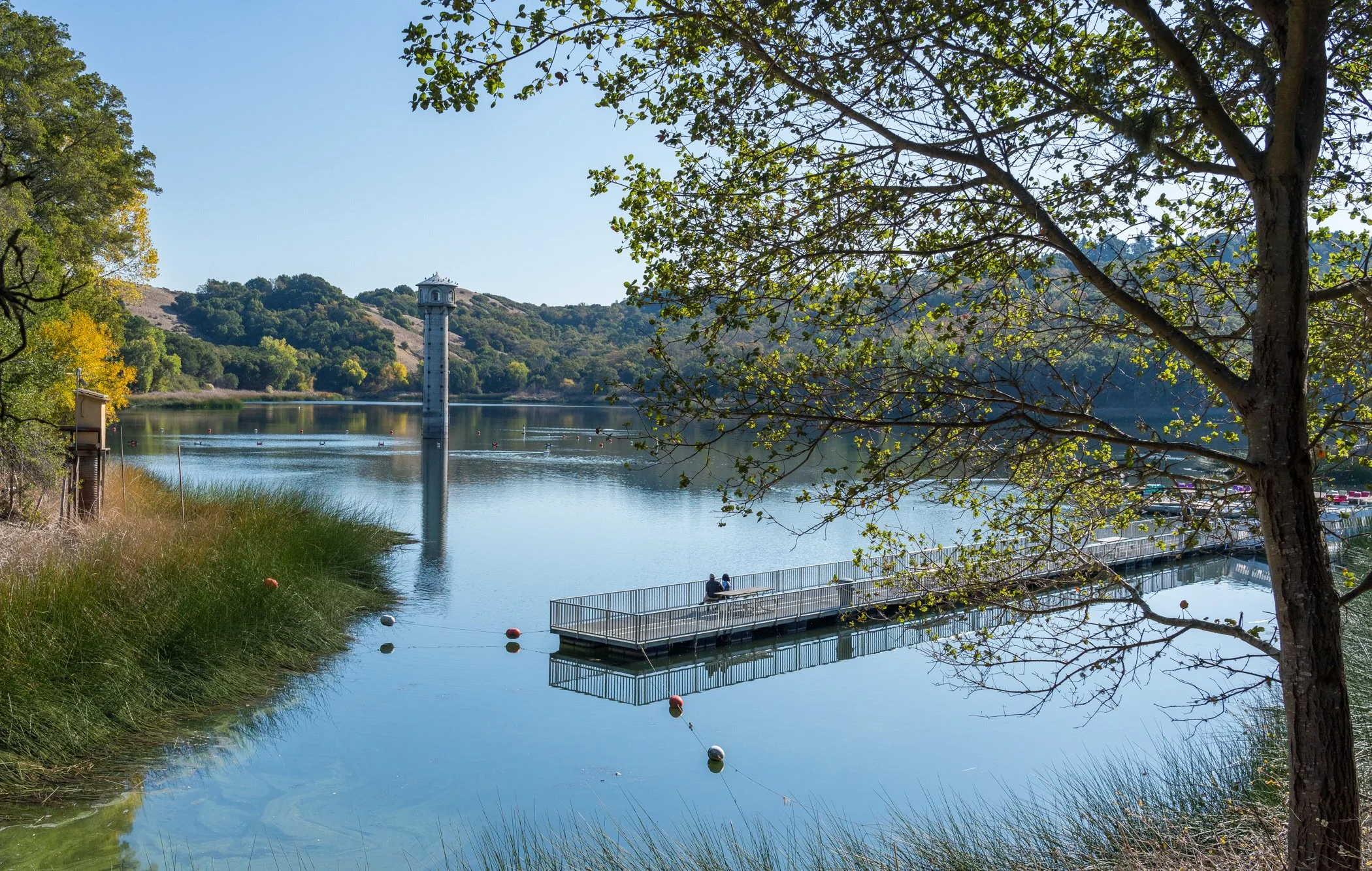 Scenic lake with a floating dock, rowing course buoys, historic water tower, and tree-covered hills