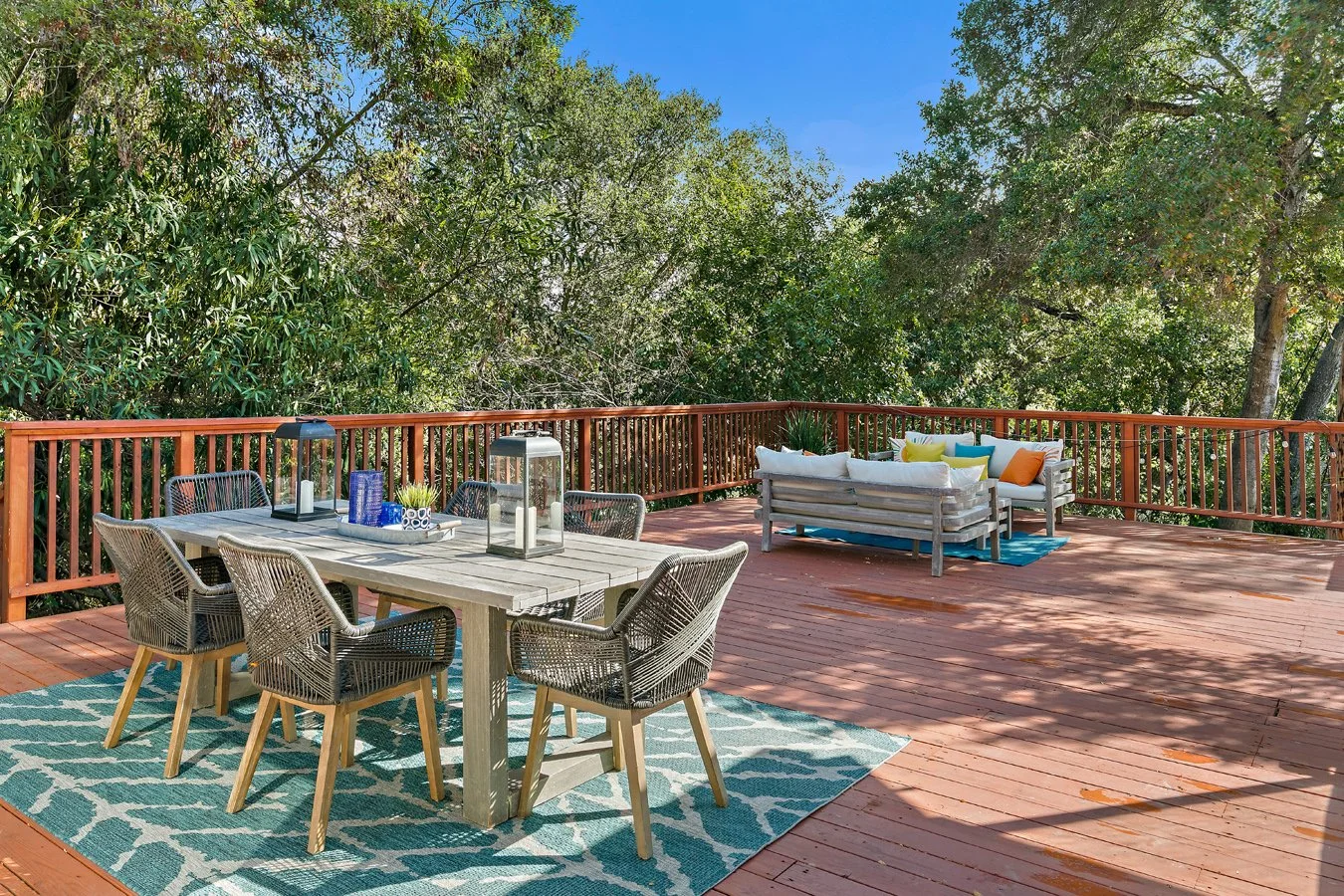 Spacious wooden backyard deck surrounded by trees, furnished with a dining table, wicker chairs, outdoor sofa, and colorful cushions.