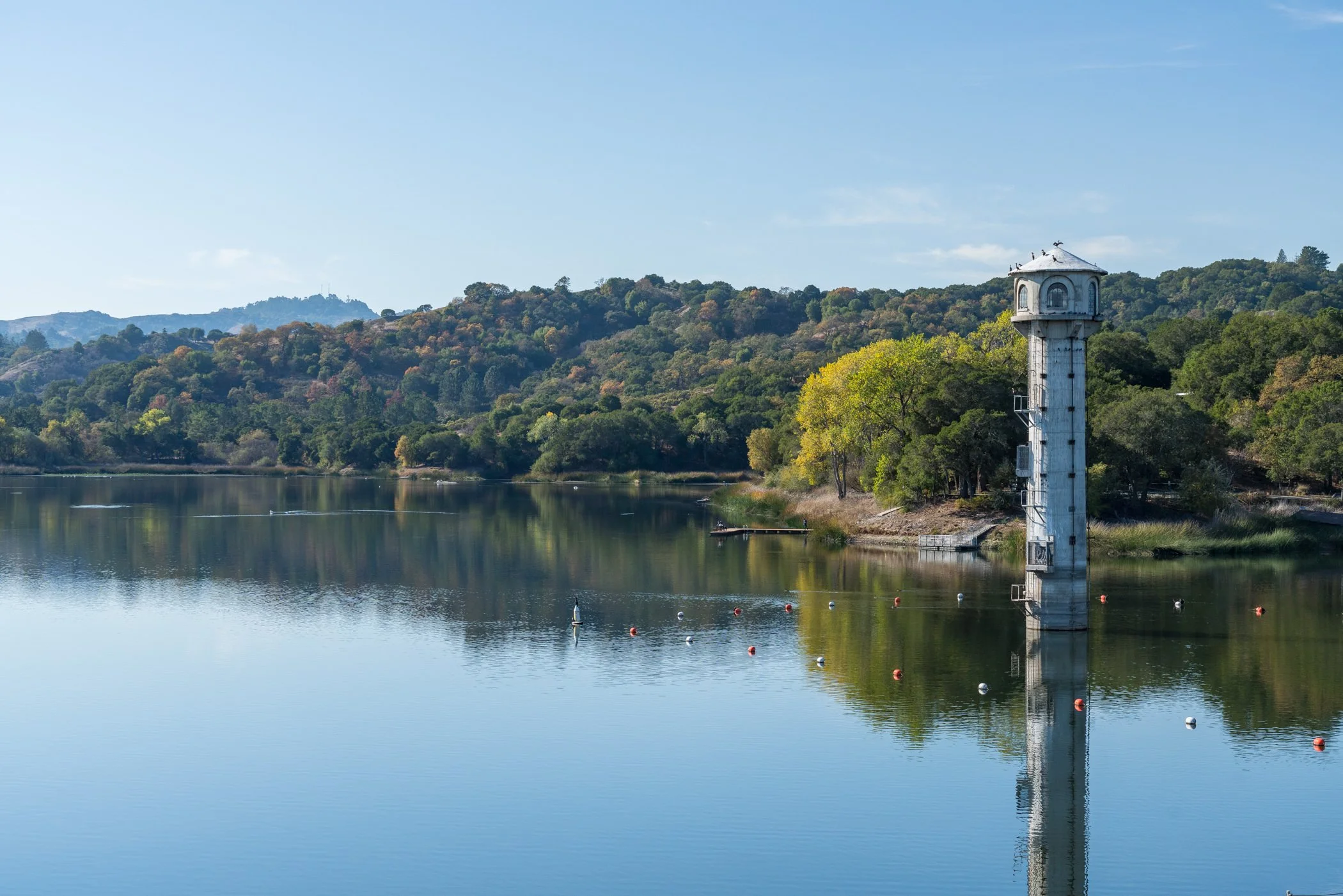 Lakeside water tower reflected in a calm reservoir, with wooded hills and buoys under a clear blue sky