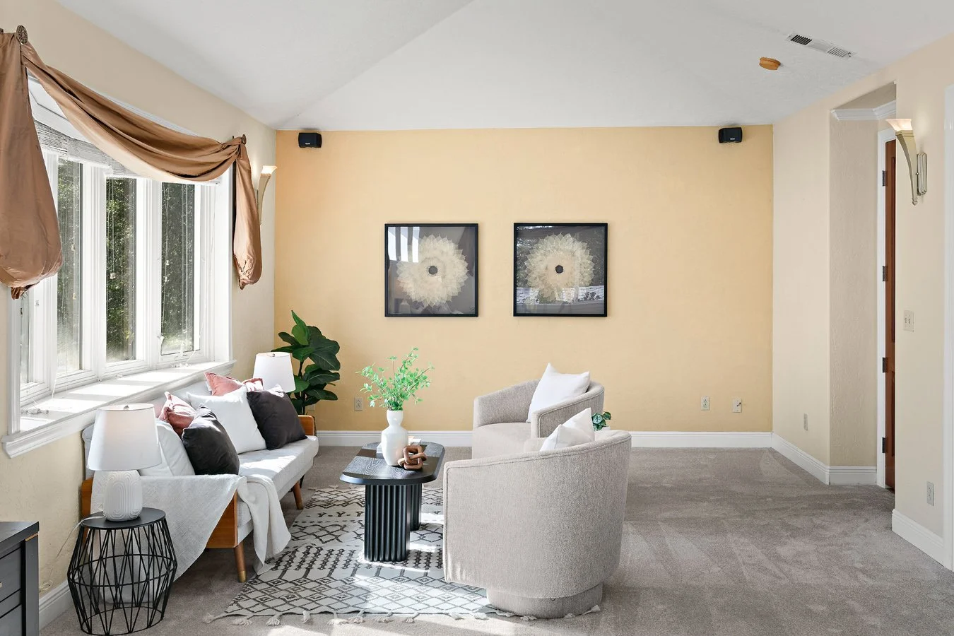 Bright living room with a white sofa, two armchairs, coffee table, patterned rug, large bay window, plants, and floral wall art.
