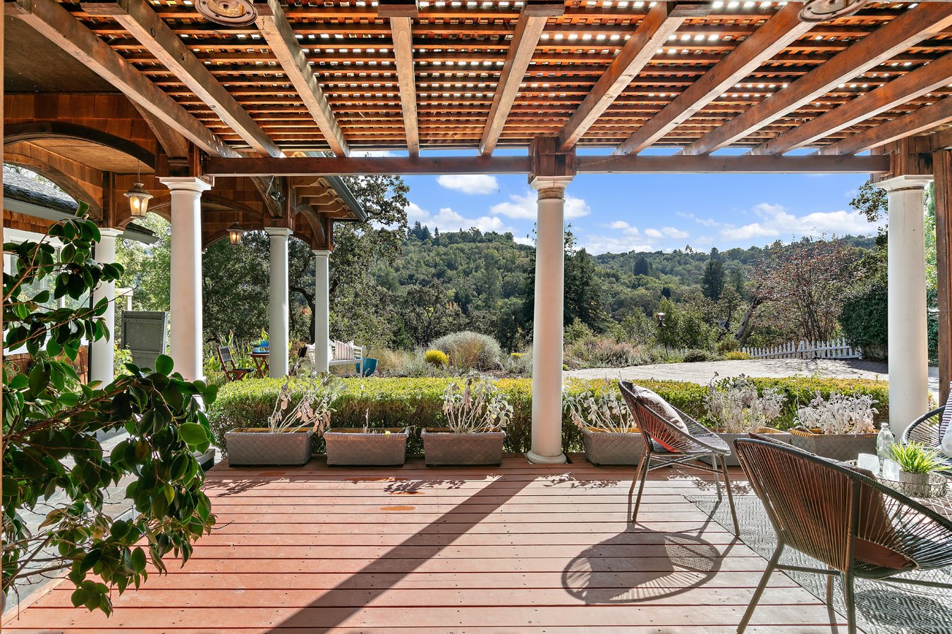 Covered wooden patio with white columns, outdoor chairs, potted plants, and wooded hillside views