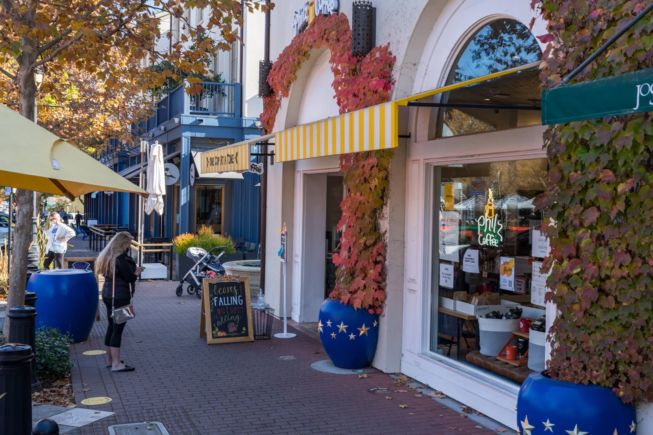 Autumn street scene with a coffee shop, striped awnings, outdoor seating, colorful ivy, and pedestrians on a brick sidewalk