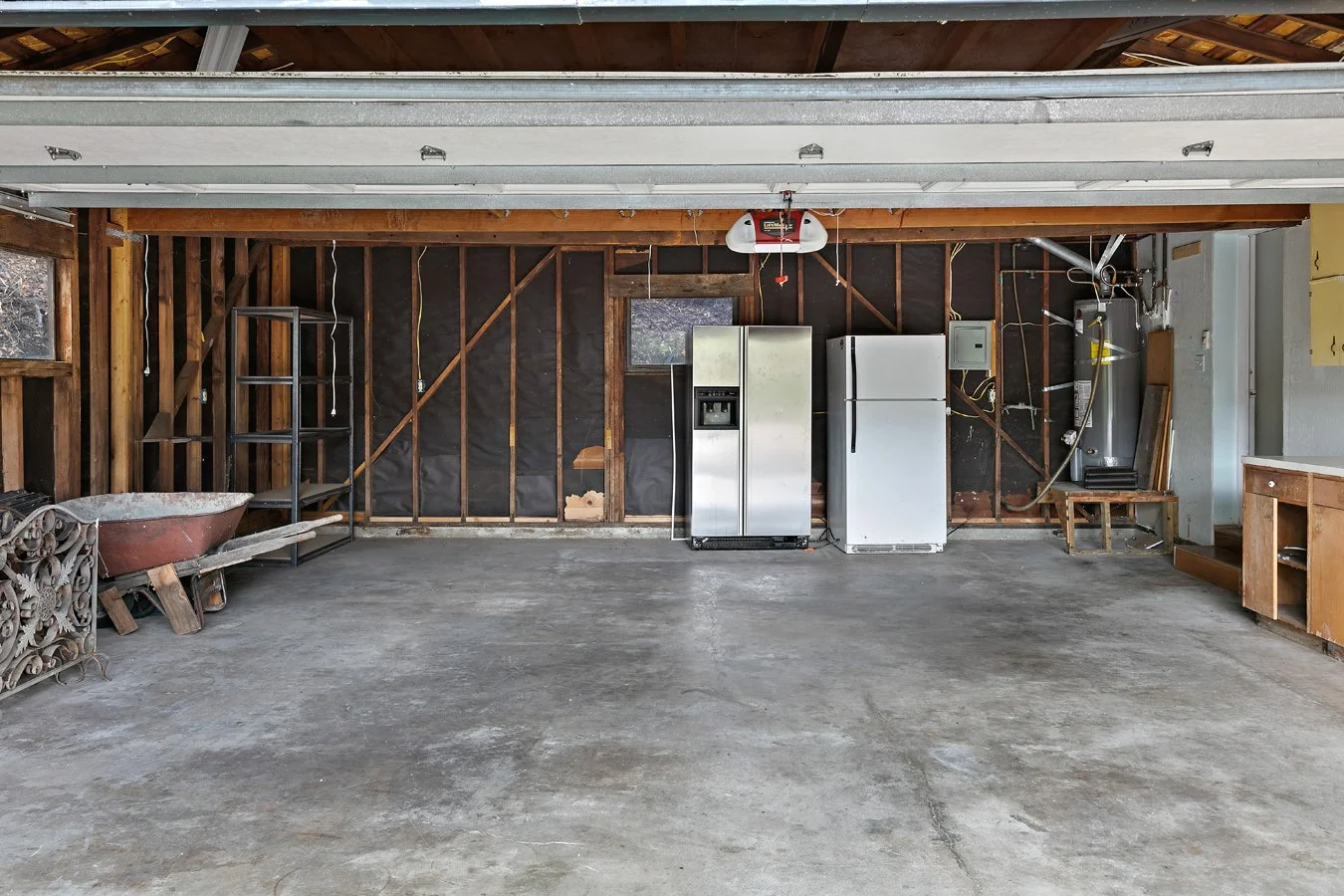 Open residential garage with exposed framing, concrete floor, two refrigerators, water heater, shelving, workbench, and wheelbarrow