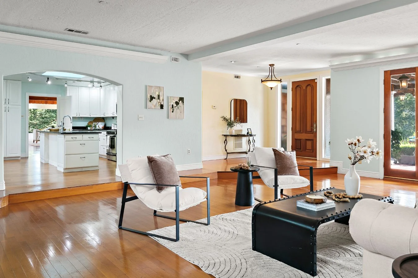 Bright open-concept living room with white seating, black coffee table, hardwood floors, and adjoining white kitchen