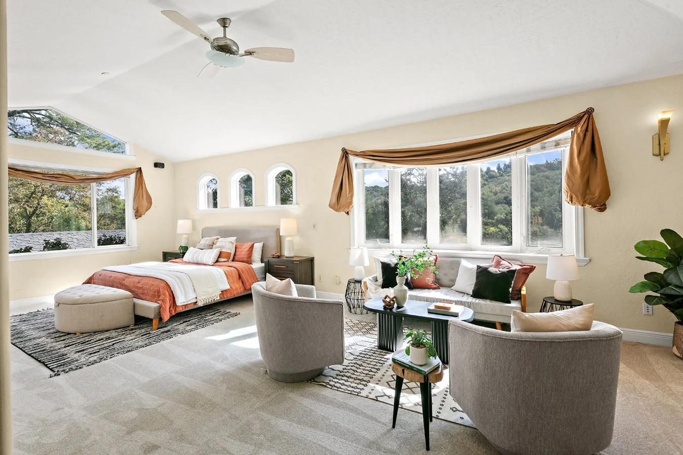 Bright, spacious bedroom and sitting area with a four-poster-style bed, sofa, armchairs, bay windows, ceiling fan, and neutral decor.