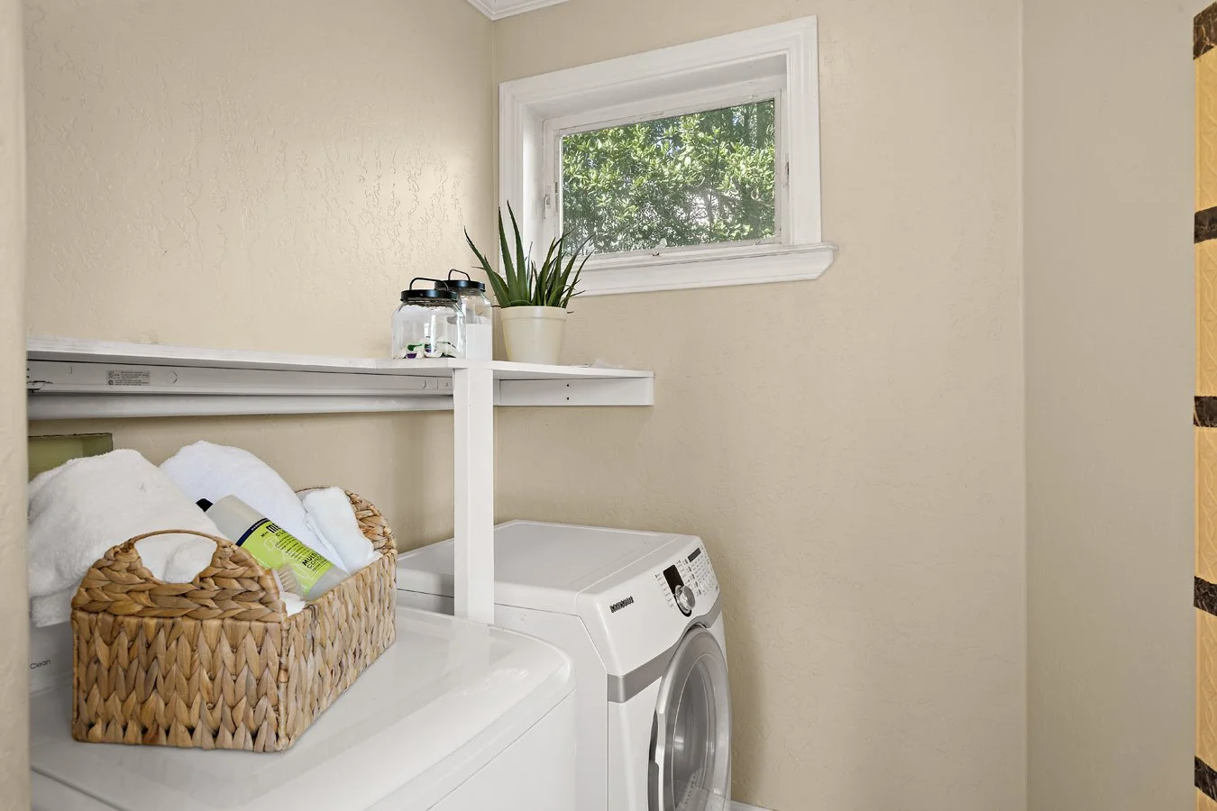 Bright laundry room with washer and dryer, woven basket of towels, detergent, potted plant, and window.