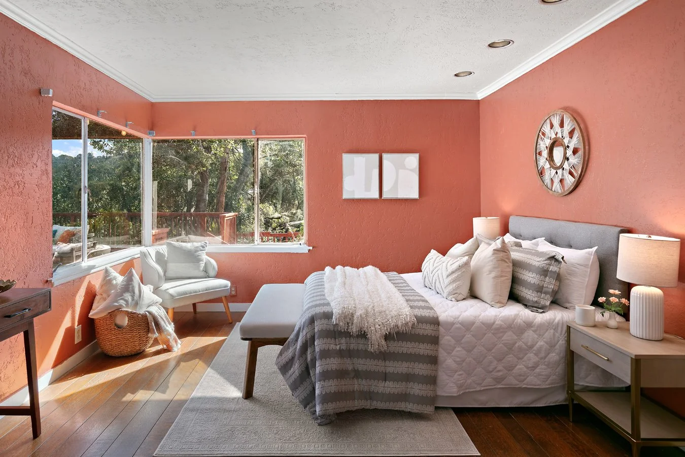 Sunlit bedroom with coral walls, a neatly made bed, large windows overlooking trees, a bedside table, and cozy seating.
