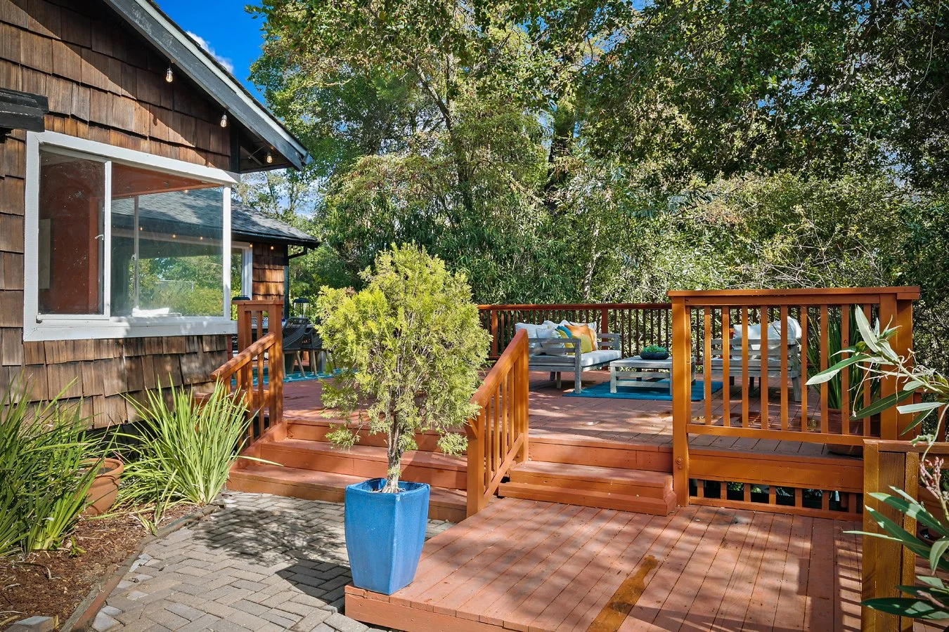 Sunlit wooden deck with outdoor seating, railings, potted plants, and a shingled cottage surrounded by trees