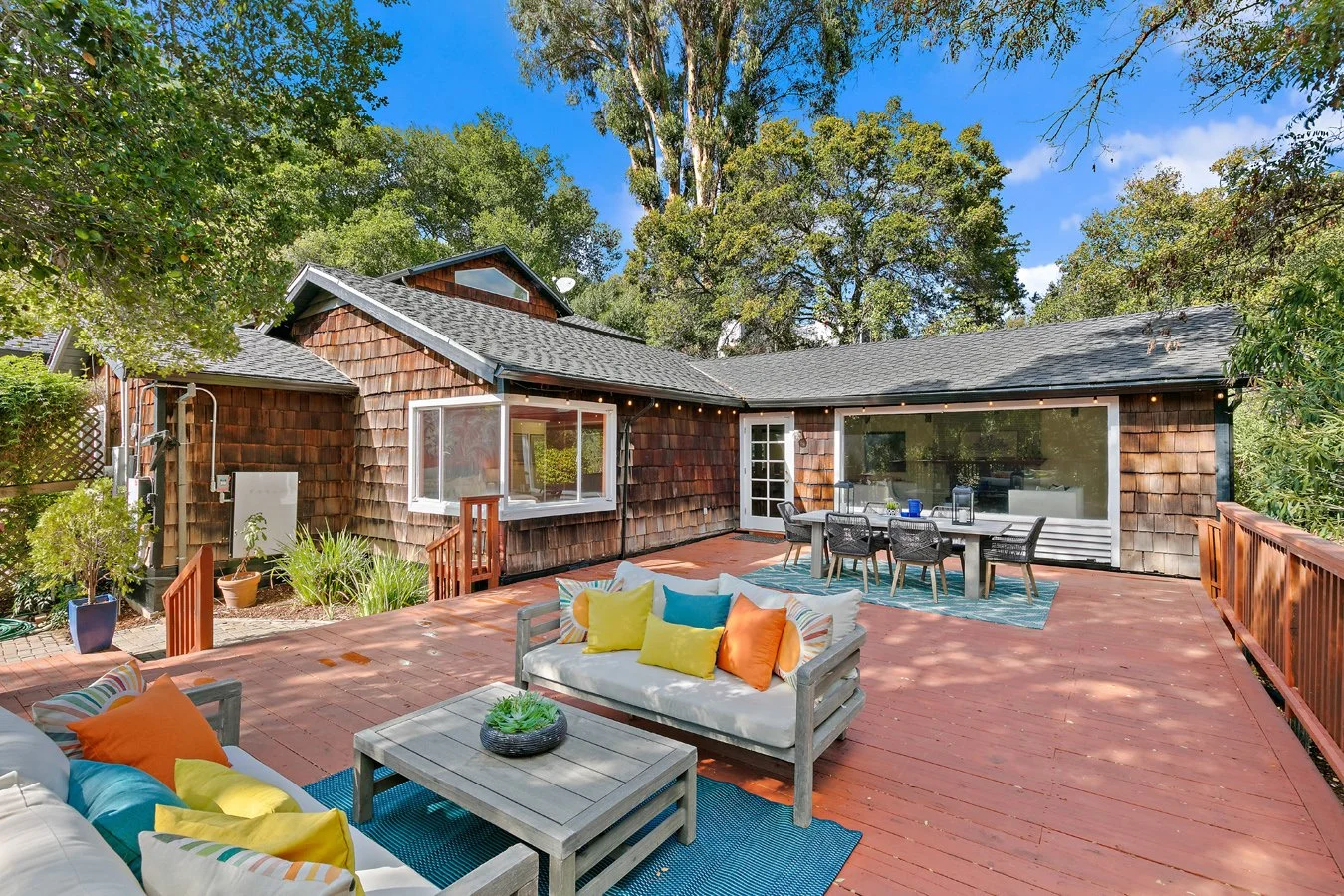 Spacious cedar-shingle home with a furnished redwood deck, outdoor dining area, colorful seating, and surrounding trees under a blue sky