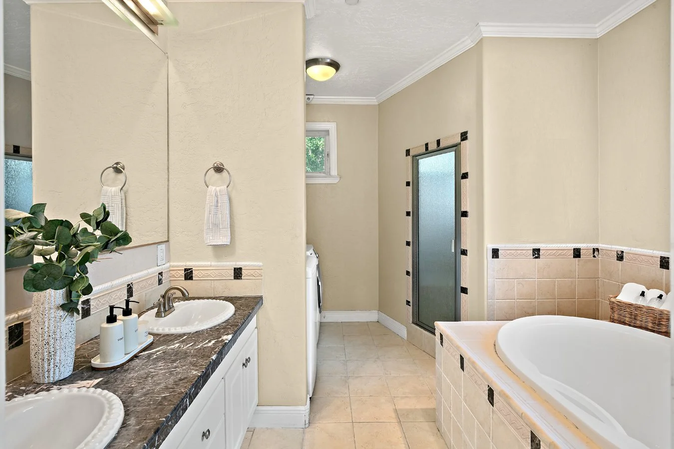 Bright beige bathroom with double-sink vanity, marble countertop, large mirror, tiled bathtub, and frosted-glass shower door