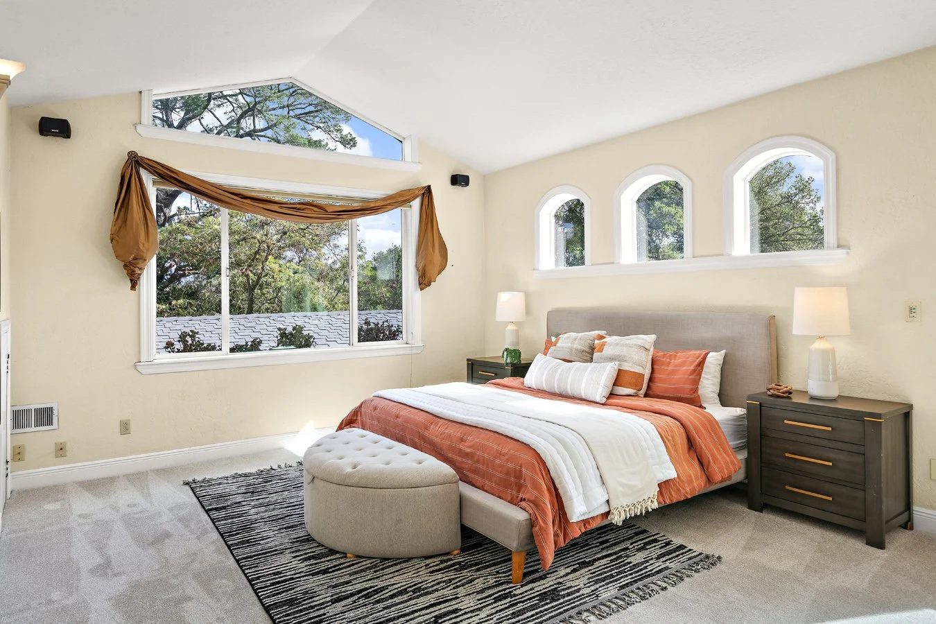 Bright bedroom with a queen bed, orange and white bedding, upholstered bench, black dresser, lamps, area rug, and large windows overlooking trees