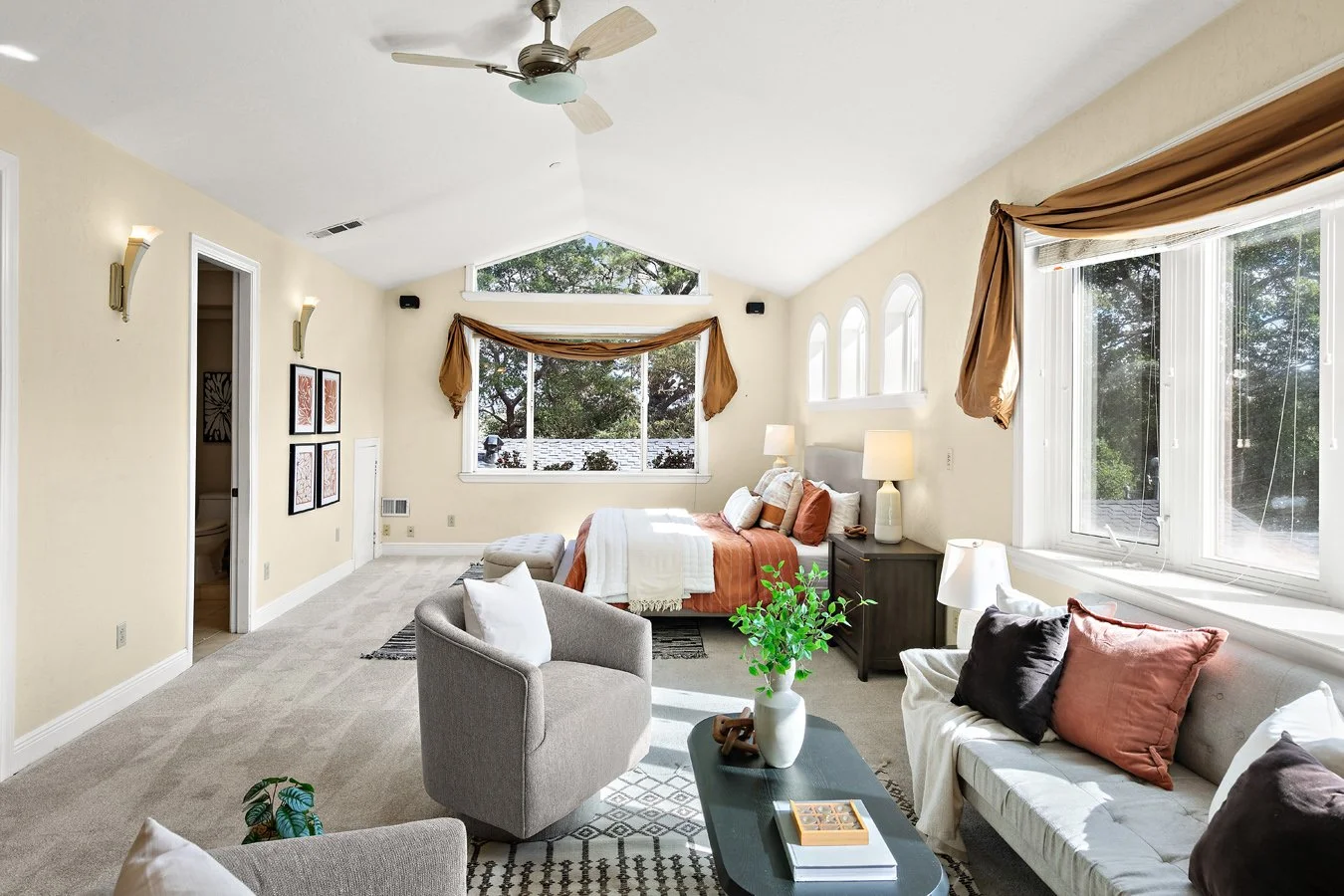 Bright bedroom sitting area with a bed, sofa, armchair, coffee table, ceiling fan, and large windows.