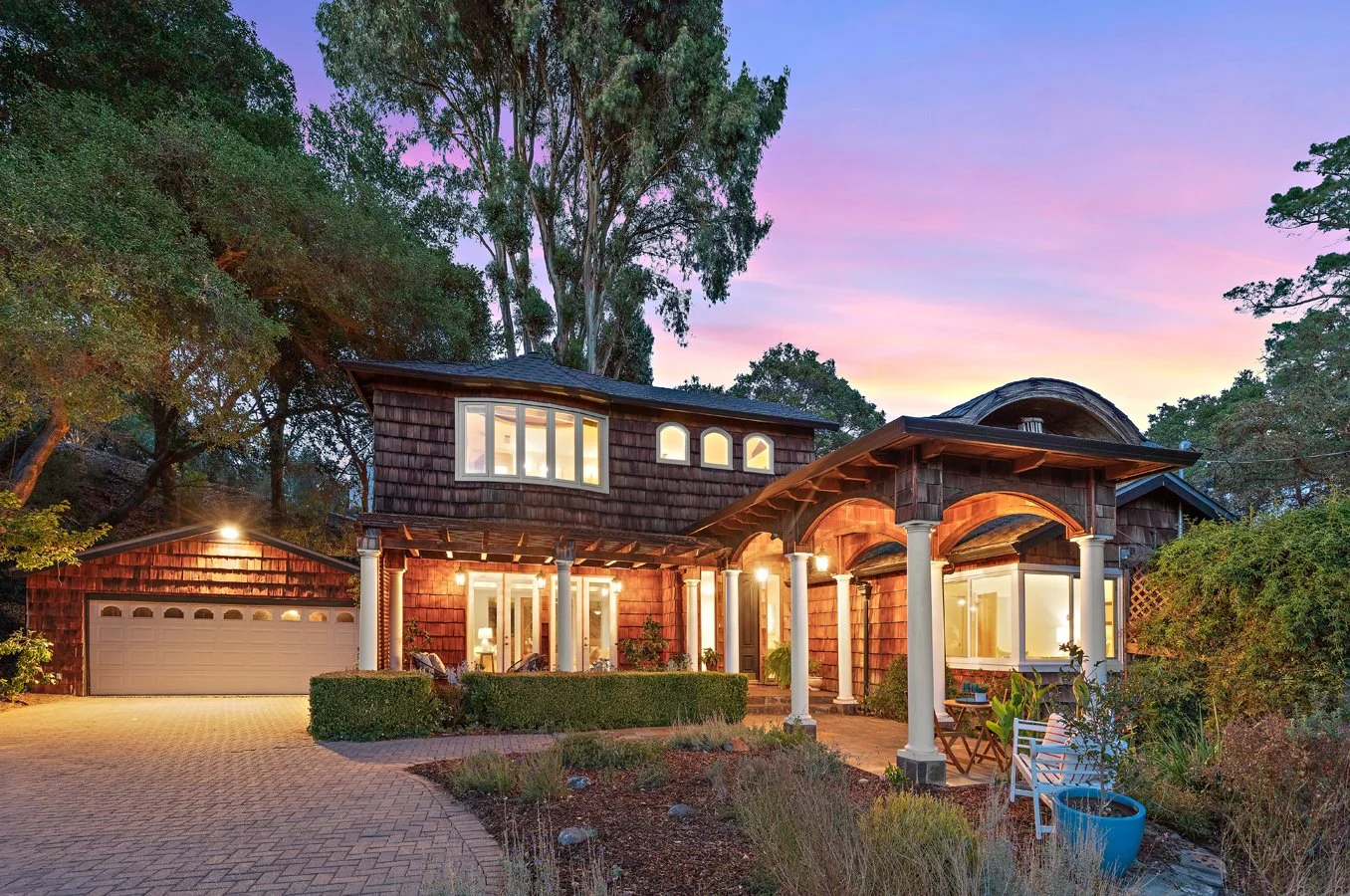 Large shingle-style house with arched porch, white columns, attached garage, landscaped courtyard, and sunset sky