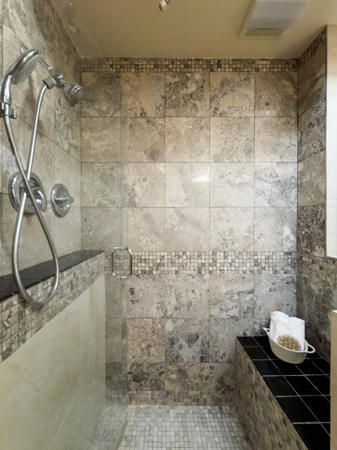 Walk-in shower with beige stone tile walls, mosaic accents, handheld showerhead, built-in bench, and tiled floor