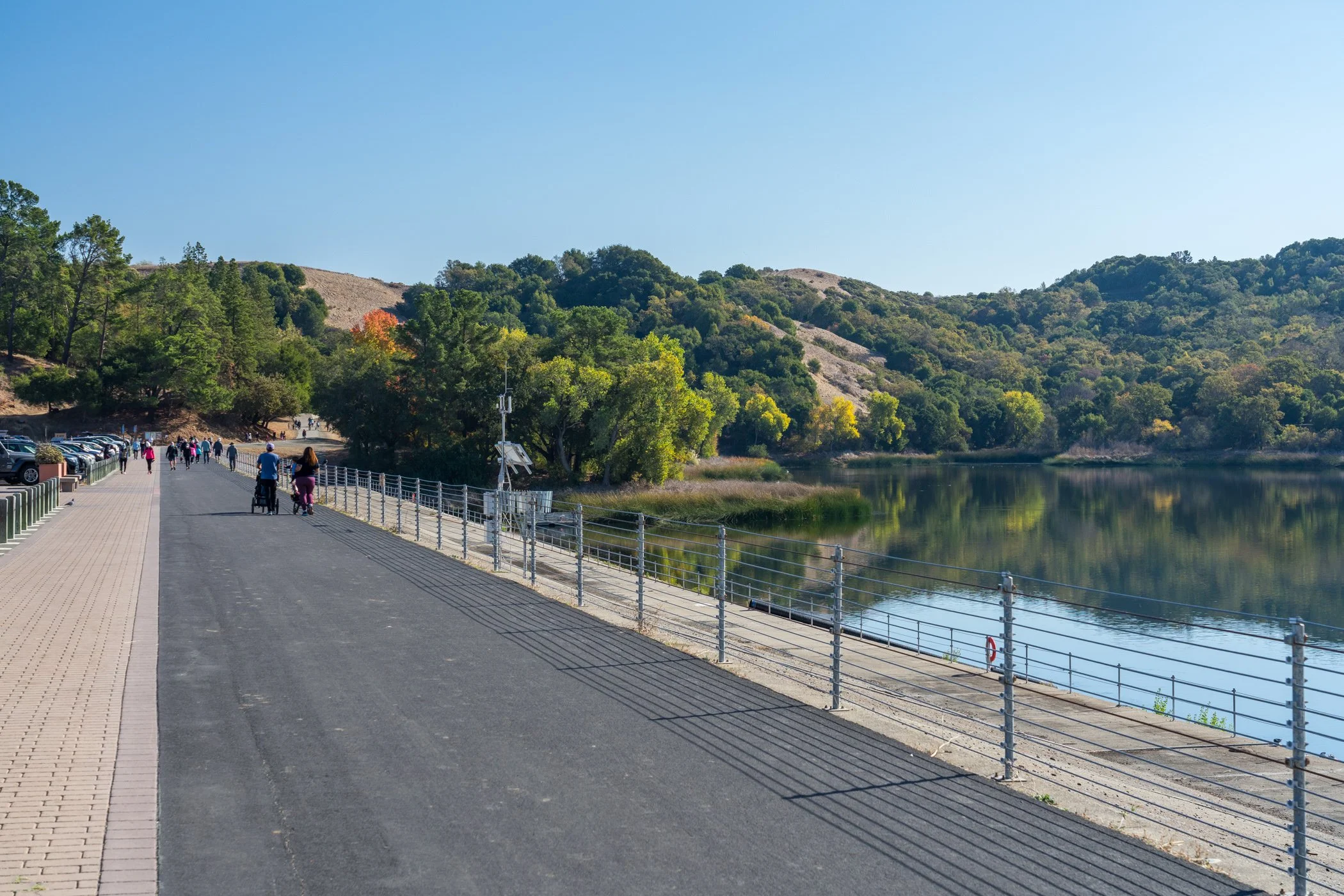 People walking and cycling along a lakeside path surrounded by autumn trees and rolling hills